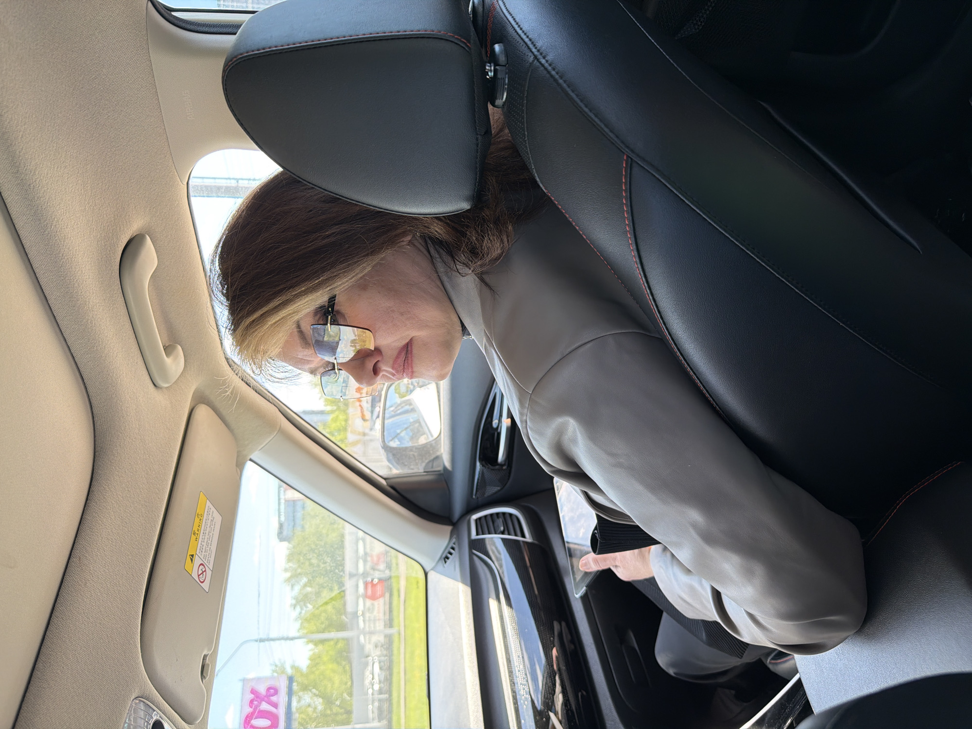 A person with short brown hair and wearing glasses and a light-colored jacket is sitting in the passenger seat of a car. The car has black leather seats with red stitching and a beige interior. Through the windshield, a street scene is visible with trees and a store with a sign featuring a percentage symbol. It is daytime. The location appears to be in the city of Krasnoyarsk, Russia.