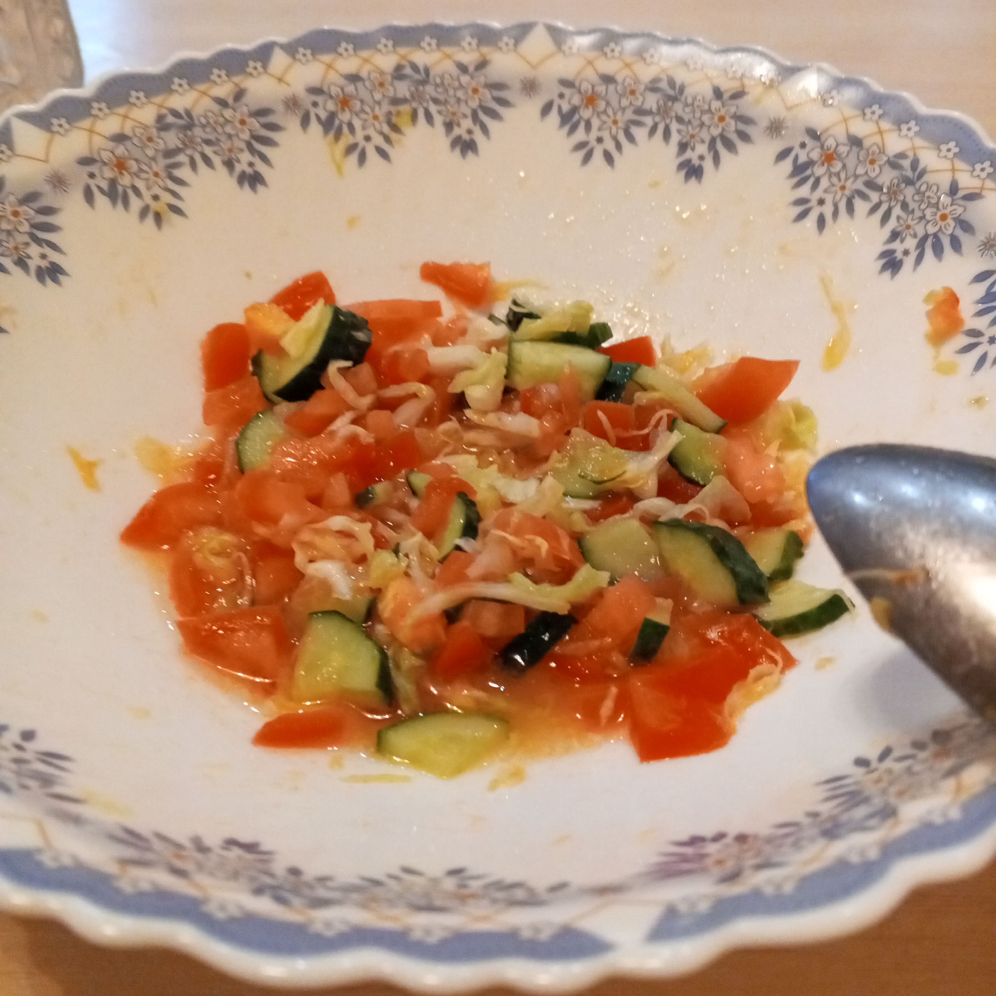 A close-up, top-down shot shows a white, scalloped bowl with a blue and white floral pattern along its rim. The bowl contains a salad made of chopped tomatoes and cucumbers, mixed with what appears to be shredded lettuce or cabbage and a light, orange-colored dressing. A metal spoon is visible on the right side of the bowl, partially obscuring the salad. The background consists of a light-colored tabletop. There is no visible indication of the location being Prokopyevsk, Russia.