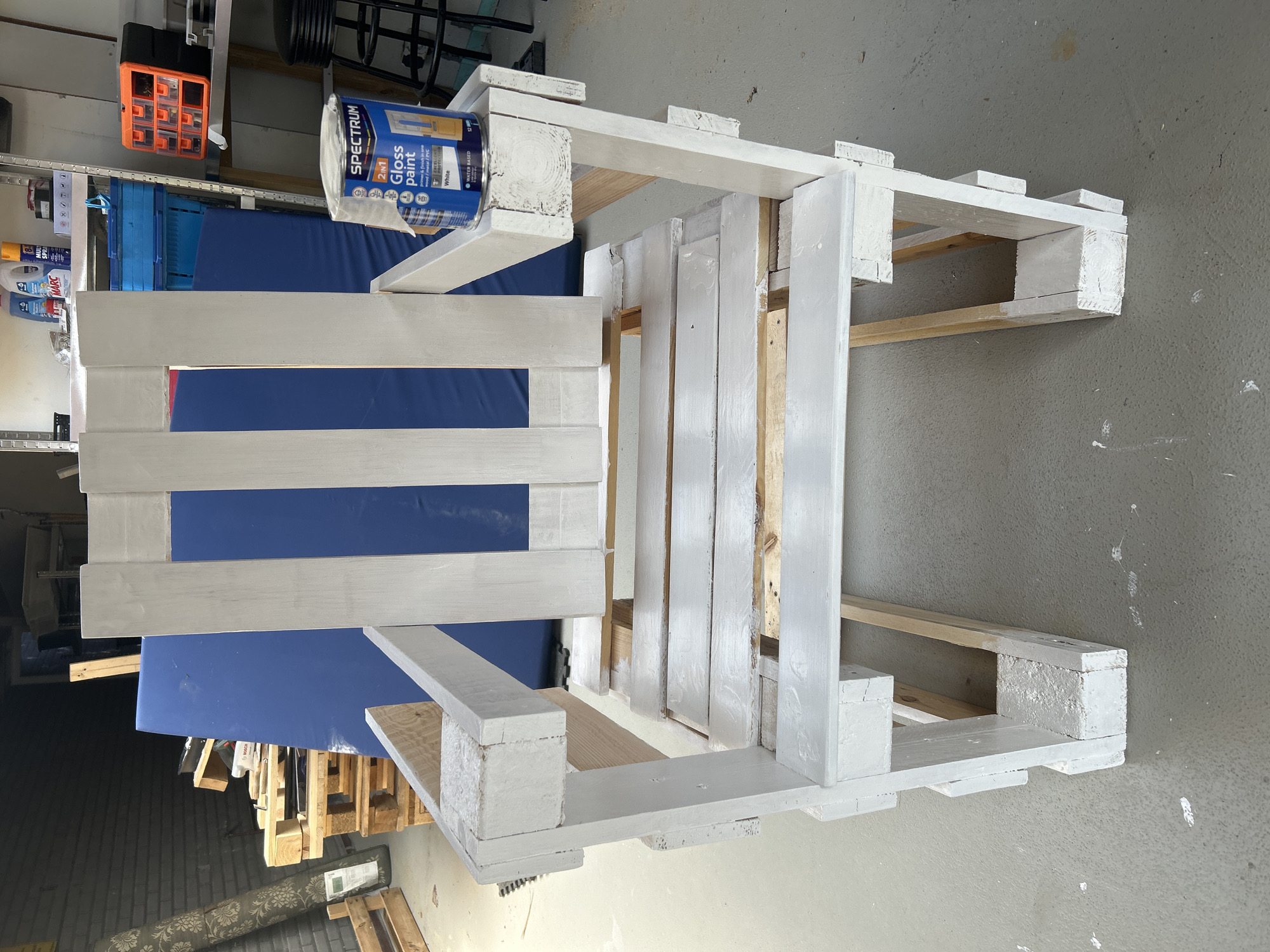 The image shows a white-painted chair made from wooden pallets, placed on a concrete floor in what appears to be a garage or workshop. The chair is in the foreground and is the primary subject. A can of "Spectrum Gloss paint" in white is visible on the armrest of the chair. In the background, there are shelves with various items, including bins and cleaning supplies, suggesting a storage or workspace. A large blue sheet or mat is leaning against the wall behind the chair. The overall scene is cluttered and industrial, consistent with a DIY project or a workspace. The image does not provide any information to suggest this location is in Lisse, Netherlands.