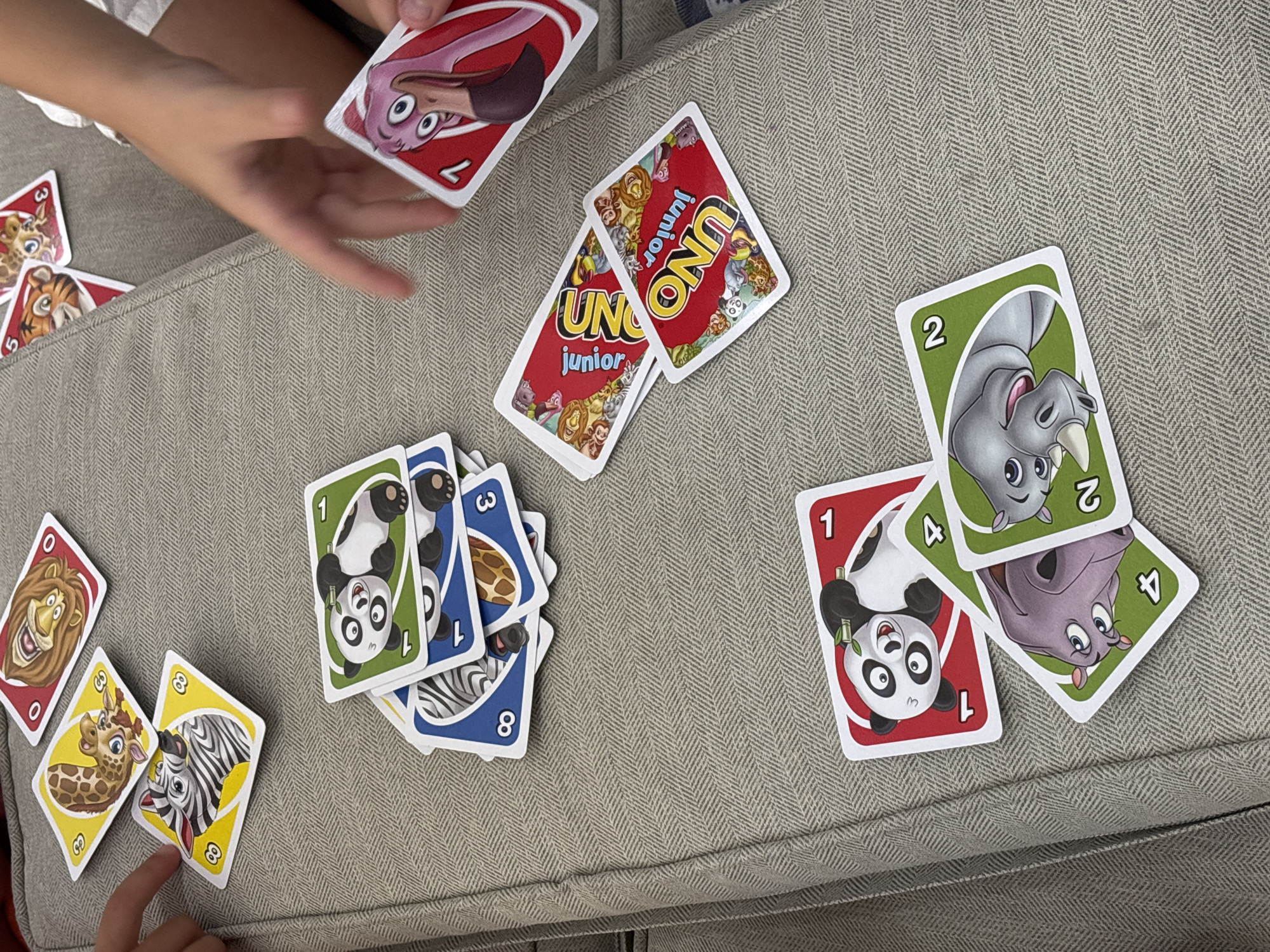 The image shows a close-up, overhead view of a game of UNO Junior being played by children on a textured, grey fabric surface, possibly an outdoor couch or seating area. Several UNO cards are scattered across the surface. In the upper portion of the image, a hand is reaching towards a red UNO card featuring a pink flamingo with the number 1. To the left, another hand is pointing towards a yellow UNO card with a zebra and the number 8. Other visible cards include a red card with a roaring lion (number 0), a yellow card with a giraffe (number 3), a stack of blue cards, a green card with a panda (number 1), a red card with a panda (number 1), a green card with a purple creature (number 4), and a green card with a rhino (number 2). Two UNO Junior cards, red with the UNO Junior logo and animal illustrations, are also visible. The lighting suggests it is daytime, and the overall scene is informal and recreational. The location is not specified in the image.