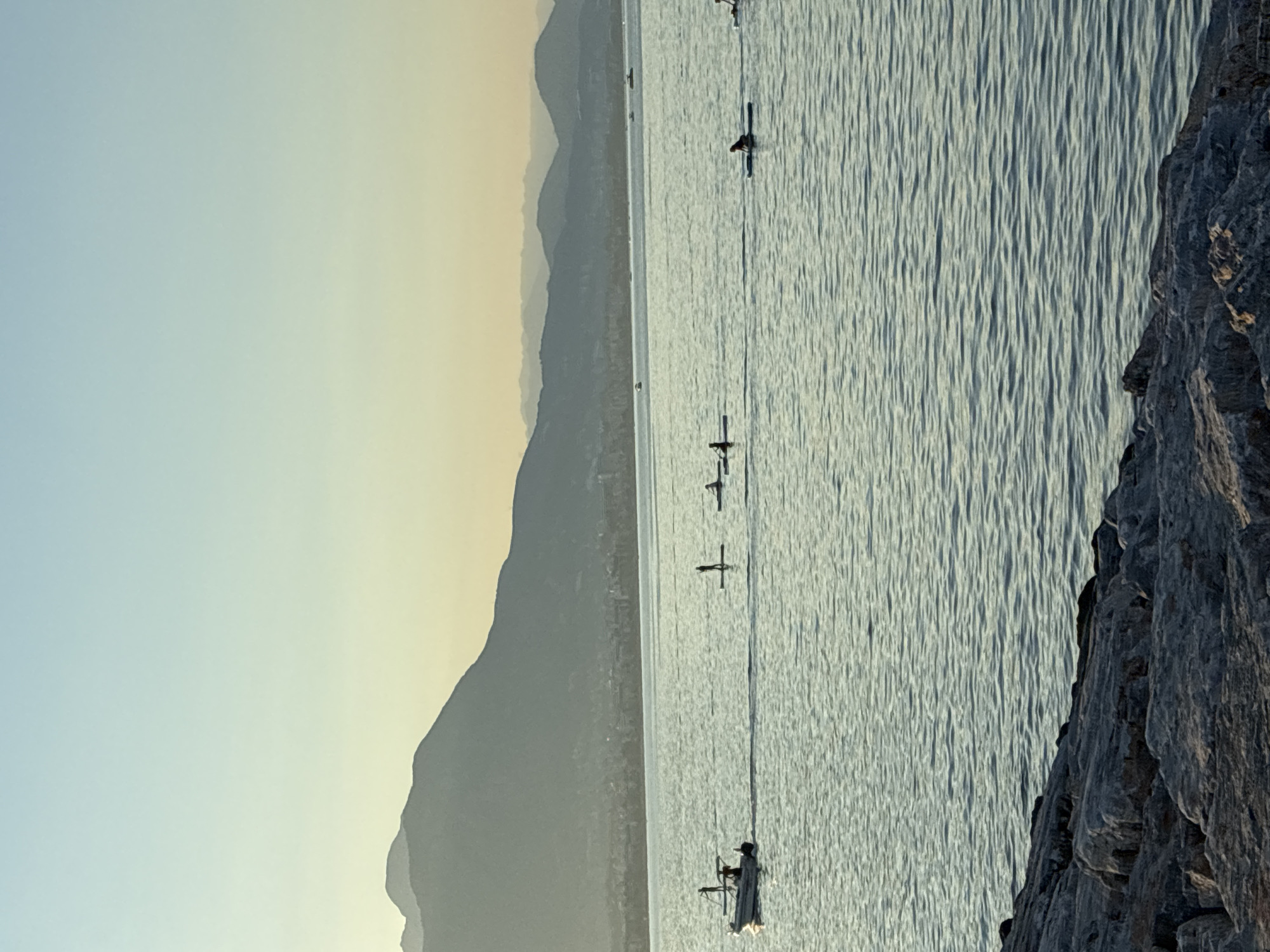 The image depicts a serene seascape with a rocky shoreline in the foreground. In the middle ground, several individuals are engaged in stand-up paddleboarding on calm, rippling water. A small motorboat is also visible, moving from left to right. In the background, a distant city skyline stretches along the coast, backed by layered mountains fading into a hazy sky. The overall scene suggests a peaceful, late afternoon or early morning setting. The location is Alanya, Turkey, evident from the distinctive mountainous landscape and coastal city profile characteristic of the region.