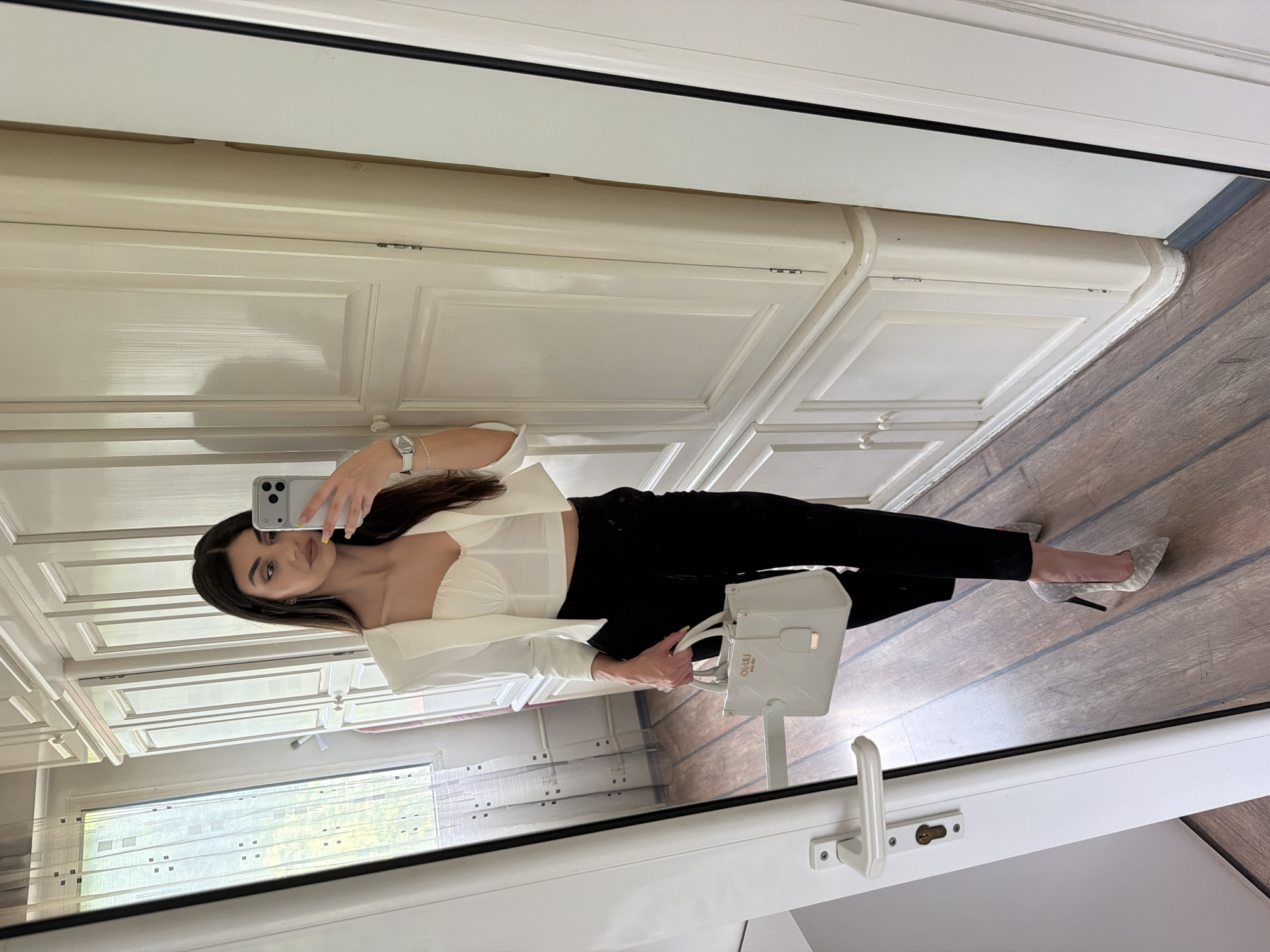 A woman is taking a mirror selfie. She is wearing a white top with long sleeves and black pants. She is holding a white handbag. She is wearing a watch on her left wrist. She is wearing high heels. The background shows a white cabinet, a window, and a wooden floor. The location in the city of Gabrovo, Bulgaria cannot be determined from the image.