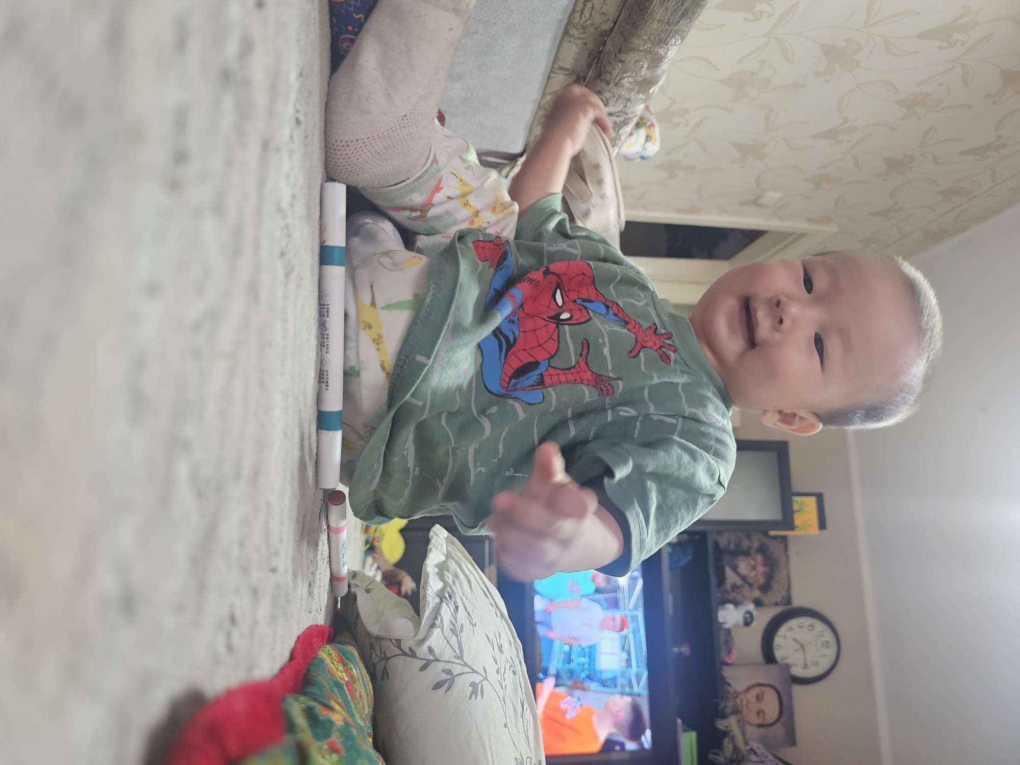 A young East Asian child is sitting on a carpeted floor in a room. The child is wearing a green t-shirt with a Spider-Man design and patterned pants. The child's left leg is visible, wearing a light-colored sock. In front of the child, on the floor, are two marker pens. In the background, a television is on, displaying an image of people. Behind the television, a clock and several framed pictures are visible on a shelf. The room appears to be a living area, with a couch and patterned wallpaper. The lighting is indoor and appears to be artificial. The child is looking towards the camera and smiling, with their right hand extended forward. The image was taken in Ak-Jol, Kyrgyzstan.