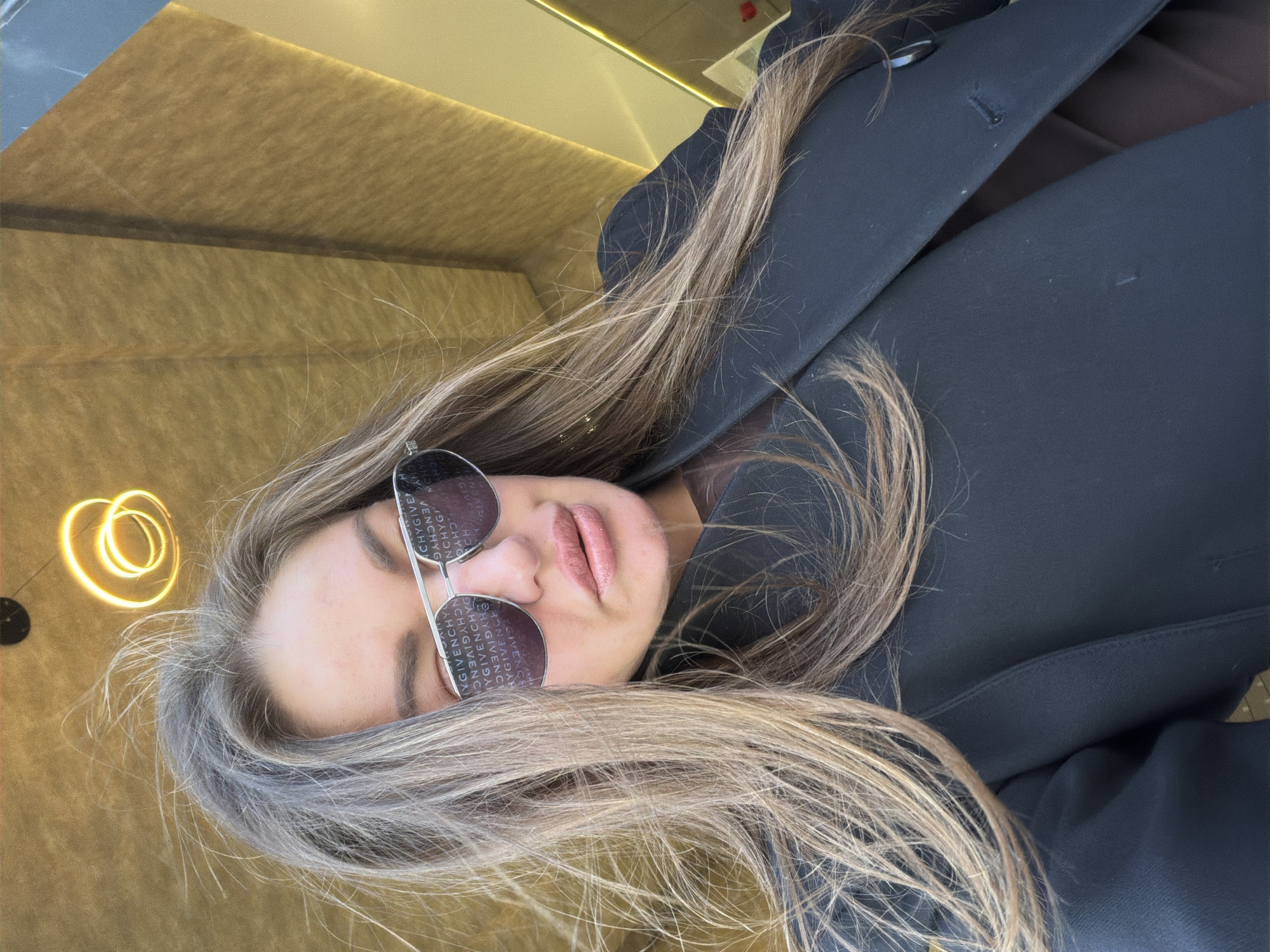 The image features a woman, seen from the chest up, positioned in an upscale indoor setting, likely a bar in Montenegro. Her head is tilted, with her long, light brown hair, highlighted with blonde streaks, flowing across the frame.
She is wearing dark, aviator-style sunglasses that prominently display the brand name "GIVENCHY" repeated across the lenses. Her lips are slightly parted, adorned with soft pink lipstick. She is dressed in a dark, possibly black or navy blue, tailored jacket or coat.
The background is dominated by textured, golden-brown wall paneling, indicating a modern and sophisticated interior. To the upper left of her head, a distinctive, circular light fixture emits a warm, golden glow, contributing to the ambiance. The overall mood conveyed is one of relaxation and style within a chic environment. The woman appears to be posing for a photograph.