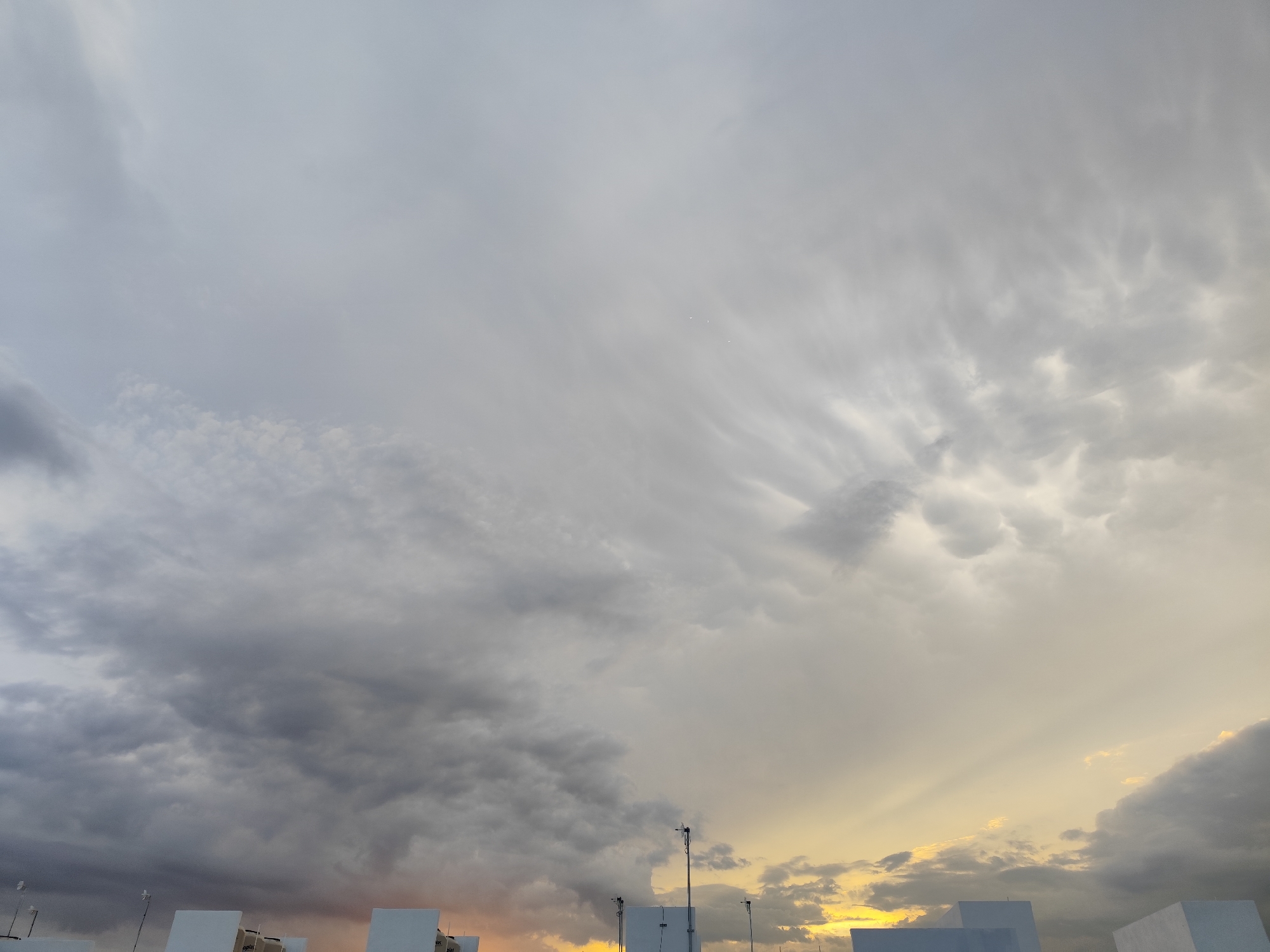 The image depicts a cloudy sky with patches of sunlight visible through the clouds. The clouds vary in density and texture, with some appearing dark and dense, while others are lighter and more scattered. There are some structures in the foreground, which seem to be rooftops or buildings with white, box-like shapes. Several metal masts or antennae protrude from the top of these structures. The scene is likely photographed at dusk or dawn, given the golden light filtering through the clouds. Without additional context, it is difficult to conclusively identify the location as Playa del Carmen, Mexico, but the style of the architecture may align with the region. There are no people visible in the image.
