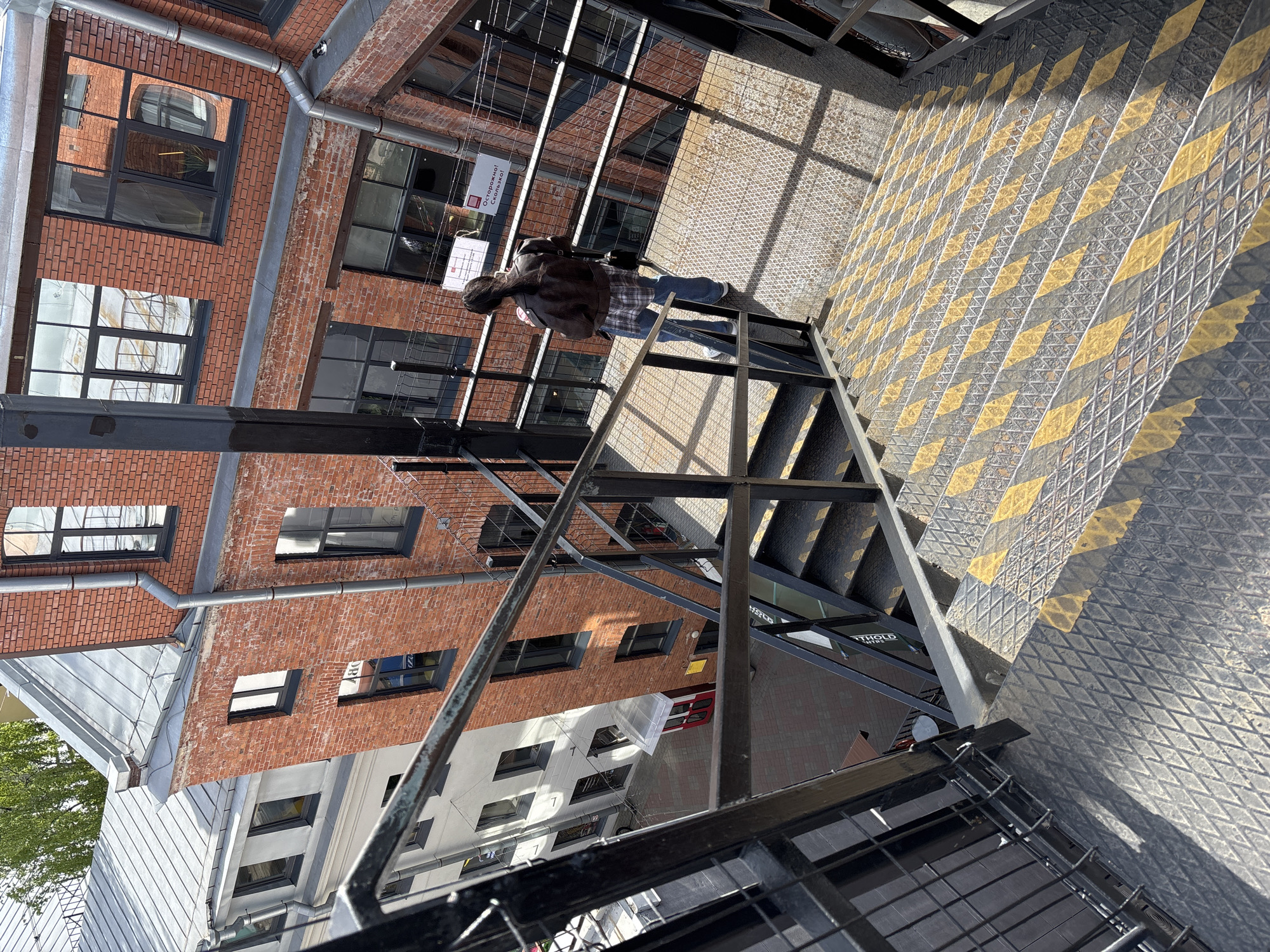 A rotated, high-angle medium shot shows a person descending a metal staircase in Saint Petersburg, Russia. The individual, dressed in a brown leather jacket and blue jeans, is midway down the stairs, with their back towards the viewer. A black backpack is on their back. The staircase has a black and yellow striped pattern on the steps and is surrounded by a metal cage. The staircase is positioned outside of an old brick building. The surrounding environment includes metal beams and a partially visible tree.