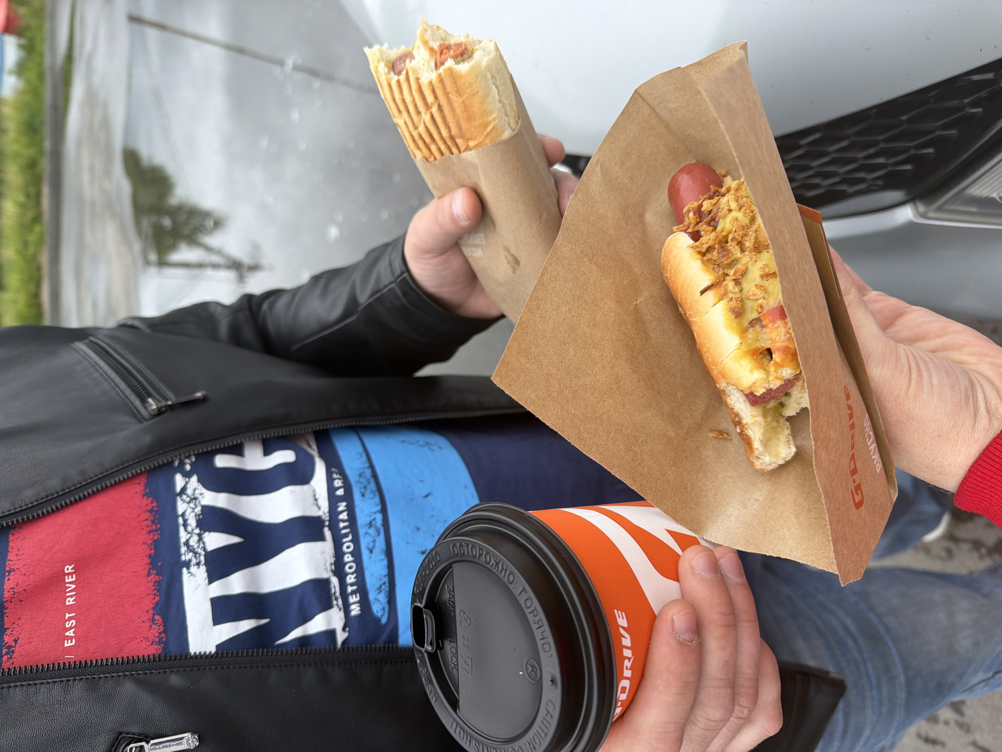A person is holding two hot dogs and a cup of coffee while seated inside or beside a vehicle. One hot dog is grilled and contained in a brown paper sleeve, while the other is topped with fried onions or a similar topping and held in a triangular paper container. The cup of coffee is orange and white with the word "DRIVE" visible on it. The person is wearing a black leather jacket with a zipper partially open, revealing a shirt with the words "EAST RIVER" and "METROPOLITAN ARFI". The setting appears to be outdoors near a car with a visible headlight and grill. There is no definitive evidence to locate the scene in Dubrovka, Russia.