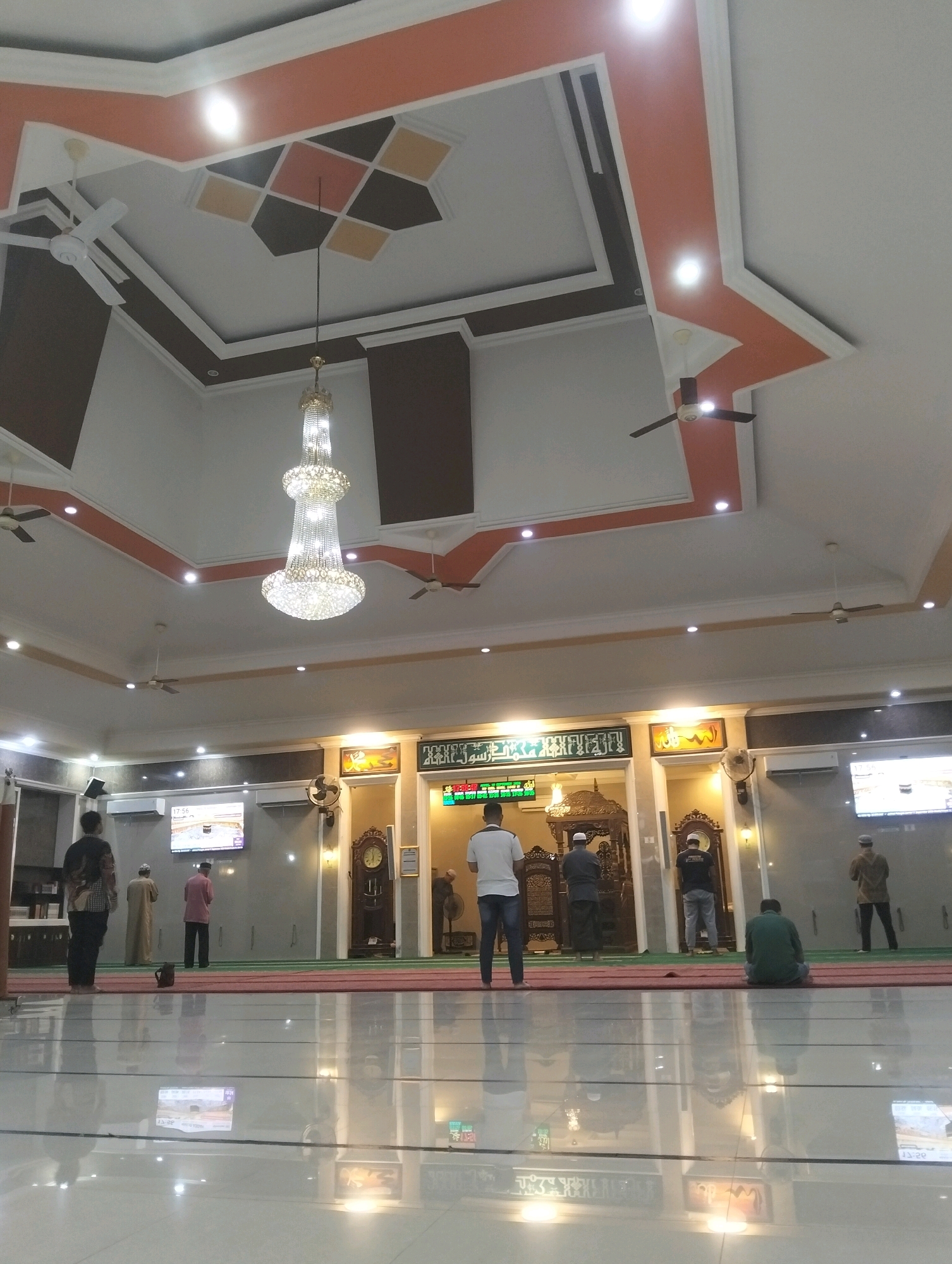 The image depicts the interior of a mosque in Palangka Raya, Indonesia, with a low-angle shot revealing the reflective floor, ceiling, and several individuals engaged in prayer or congregating. The mosque features a modern architectural style with a multi-layered ceiling adorned with geometric patterns in orange, dark brown, and white. A prominent, ornate crystal chandelier hangs from the center of the ceiling, surrounded by multiple ceiling fans and recessed lighting.
In the foreground, the highly polished, tiled floor reflects the ceiling lights and the figures above, creating a sense of depth and spaciousness. Several men are visible throughout the mosque. On the far left, a man in a patterned long-sleeved shirt and dark pants stands facing away from the camera. Further into the mosque, a man in a white shirt and blue jeans stands facing the mihrab, a semicircular niche in the wall indicating the direction of Mecca. To his right, a man in a dark grey thobe and headscarf faces the mihrab. Another man in a dark t-shirt and grey pants stands facing the mihrab, with his hands clasped. To the right, a man in a brown shirt and dark pants stands facing the mihrab. In the center right, a man in a green shirt is seated on the prayer mat, facing the mihrab.
The background of the mosque is characterized by a large, ornate wooden structure, likely the mihrab and minbar. Above the mihrab, calligraphic inscriptions and framed artwork are displayed on the wall. Large flat-screen televisions are mounted on the walls, displaying images, potentially of Islamic sites or prayer times. The walls are painted in muted tones of grey and beige, contrasting with the vibrant ceiling design. A large grandfather clock is visible on the wall to the right of the mihrab. The prayer mats are laid out in rows, with a prominent red border. The overall ambiance is one of solemnity and devotion, amplified by the grand architecture and the reflective surfaces.