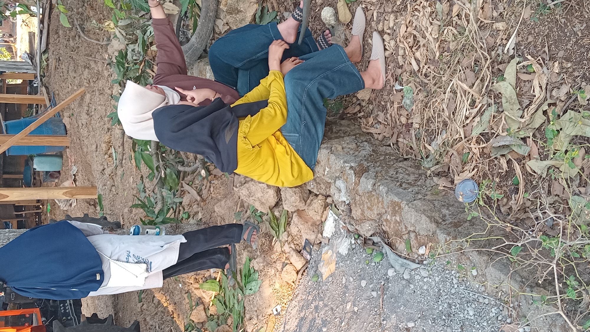 In Tinapan, Indonesia, two young women are seated on a large rock in a dry, outdoor environment. The woman on the left wears a black hijab and a bright yellow long-sleeved shirt with ruffled shoulders, paired with blue jeans. She is positioned with her legs crossed and her right hand resting on her lap. Beside her, to her right, sits another young woman wearing a light beige hijab and a brown long-sleeved top, also with blue jeans. She is pointing with her right hand towards the right side of the frame, her body angled slightly away.
The ground around them is covered with dry leaves, twigs, and some scattered rocks. To the left of the women, a portion of a grey tractor is visible, with its large black tires and orange-red body. Further in the background, a wooden structure with a slanted roof is present, and several blue and green barrels are stacked near it. A woman in a navy blue hijab and a white shirt with a small bag attached is partially visible on the far left, standing near the tractor. The overall impression is a rural or semi-rural setting with agricultural elements.