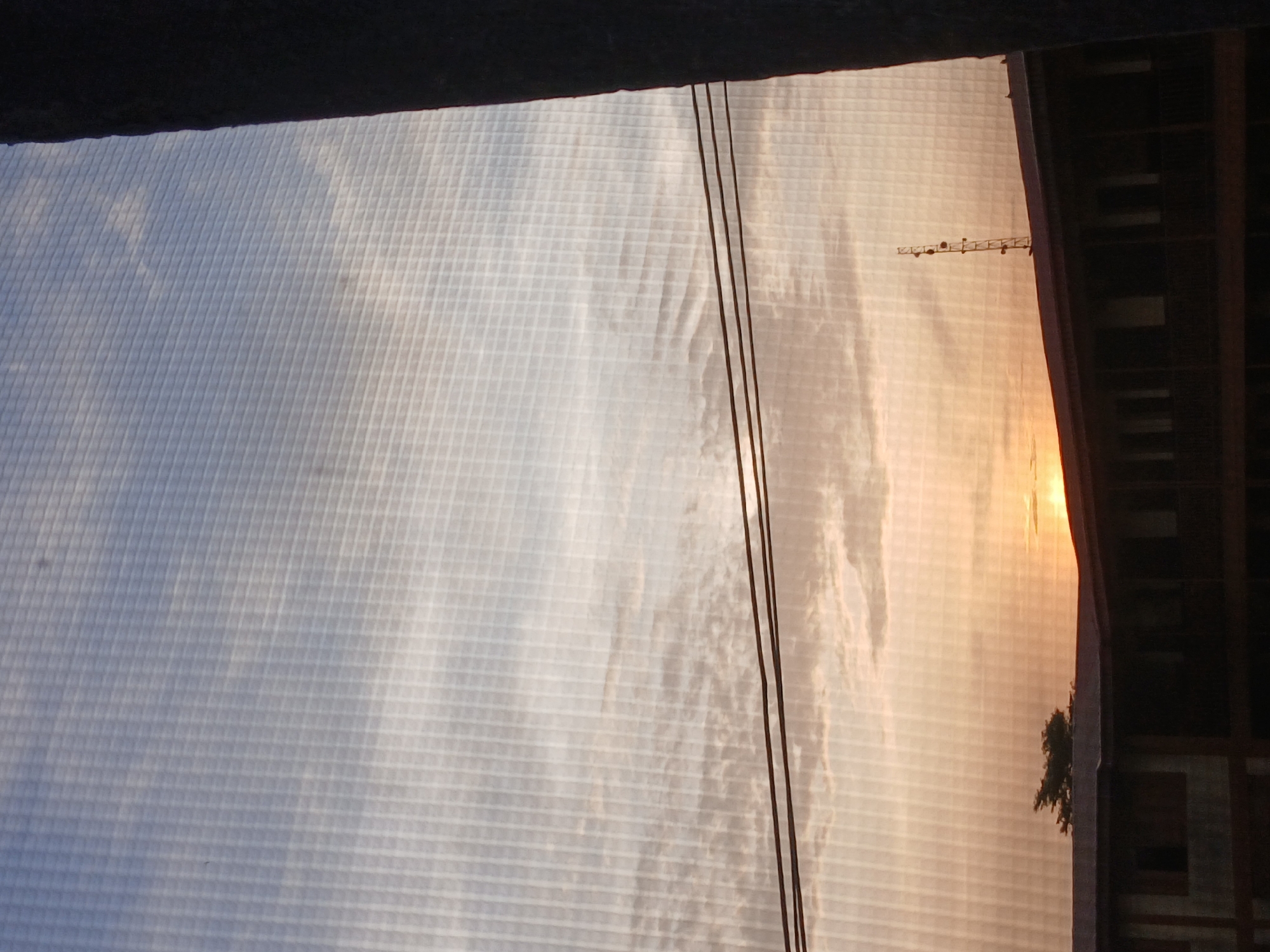 The image presents a view from behind a fine mesh screen, likely a window screen. The sky dominates the scene, exhibiting a gradient from a pale blue to a warm, orange hue, possibly indicating sunrise or sunset. Bands of white clouds stretch across the sky. Three electrical wires run vertically through the frame. The outline of a building, including windows and what appears to be an antenna, is visible on the right. A tree is partly visible beside the building. The mesh obscures a clear view of details, but the overall impression is of a cityscape at dawn or dusk. The location is identified as Lagos, Nigeria.