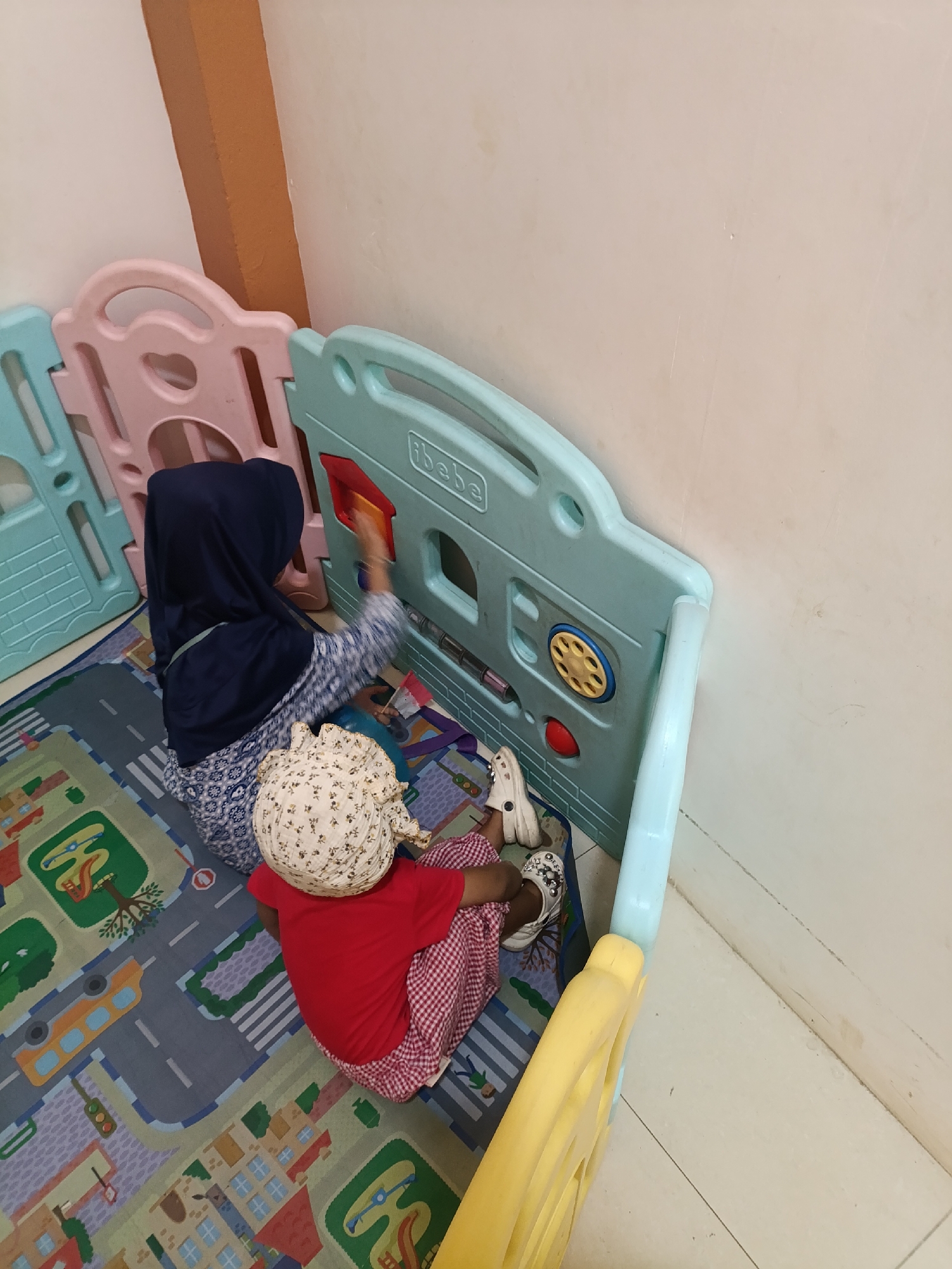 Two individuals are engaged in play within a colorful plastic playpen, situated on a mat depicting a city street scene. The playpen features panels in pink, light blue, and yellow. One individual, wearing a dark blue hijab and a blue and white patterned top, is reaching into the playpen, appearing to interact with a toy. The second individual, a child, is facing away from the camera, wearing a red shirt and a red and white checkered skirt, with a floral patterned headscarf. They are sitting on the mat, also within the playpen. The environment suggests an indoor setting, possibly a home or a play area. The image is undated and unsigned, but it is stated to be located in Jonggol, Indonesia.