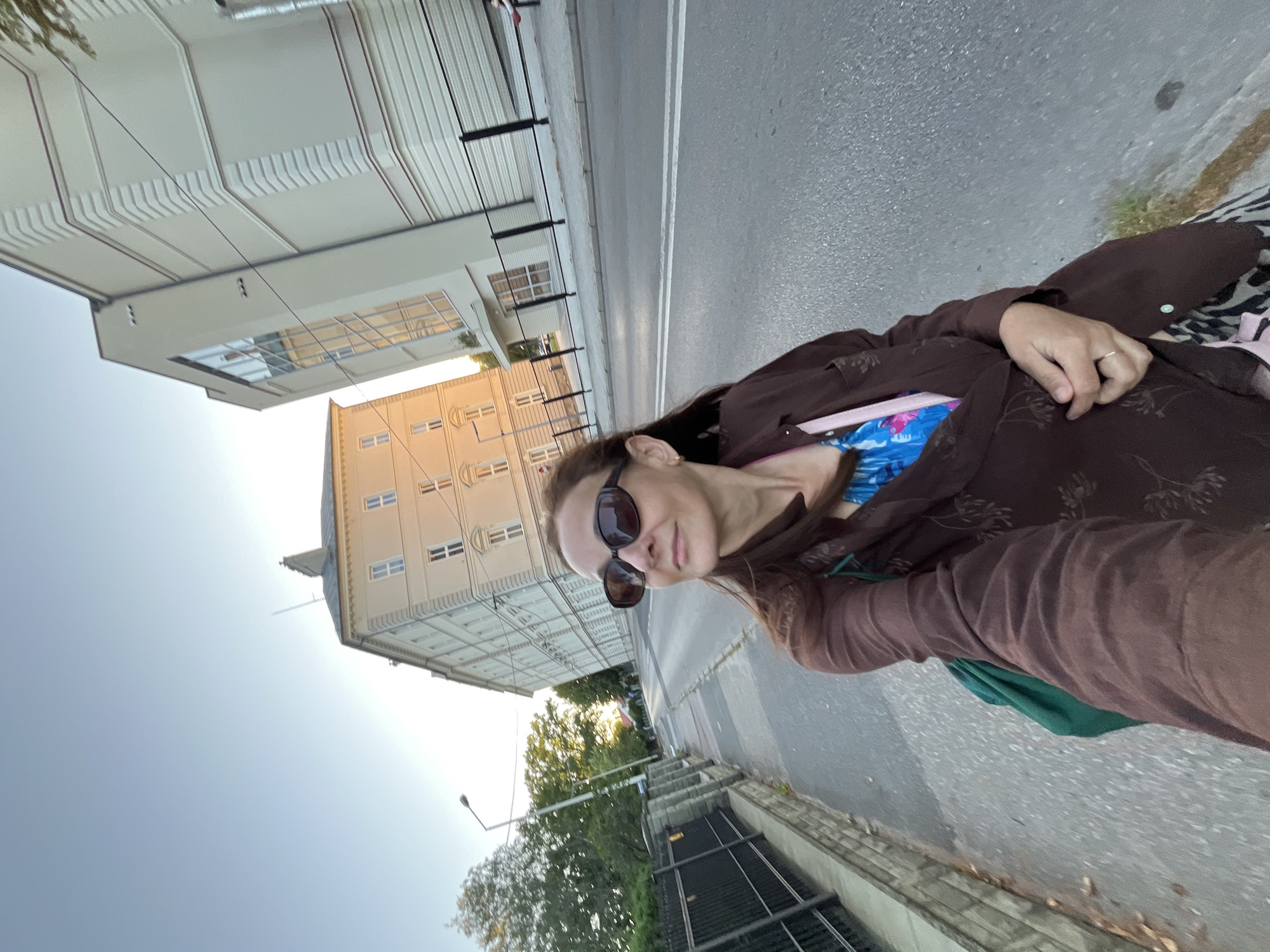 A woman with brown hair and sunglasses is taking a selfie while walking on a sidewalk in a city. She is wearing a brown jacket over a blue and pink patterned top. In the background, there are buildings and trees, and the sky is clear and blue. The street is paved with asphalt and has white lane markings. The woman is looking to her right, and her left hand is holding the front of her jacket.