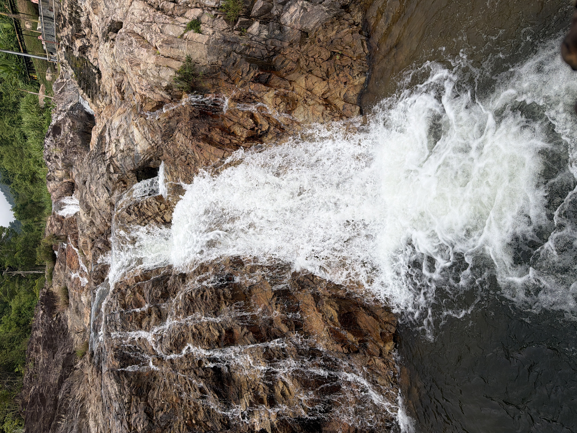 The image captures a waterfall cascading over rocky terrain in Khanh Vinh, Vietnam. The water is frothy and white as it crashes down the brown and gray rocks. Green foliage is visible on the left side of the frame. A metal barrier or walkway is partially visible at the top left corner. The water flows into a darker pool at the bottom right.