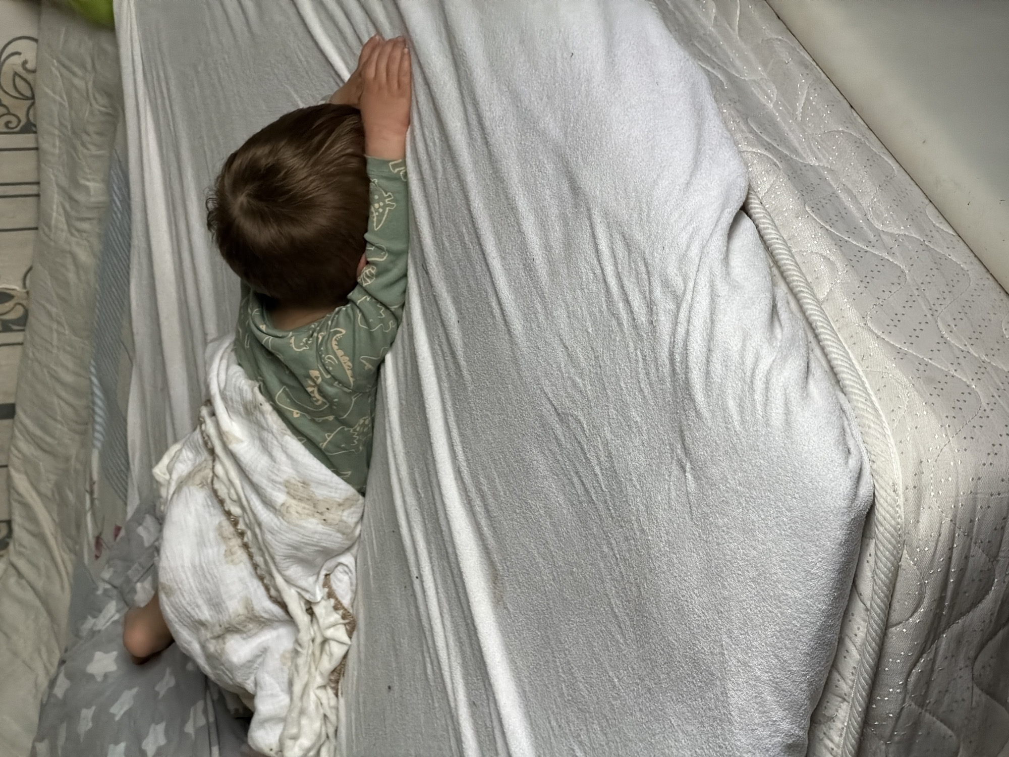 A young child with brown hair is lying on a bed. The child is wearing a green long-sleeved shirt with a dinosaur pattern. They are partially covered by a white blanket with a textured pattern. The child's hands are raised and resting on the edge of the bed. The bed appears to be in a room, possibly a bedroom, with bedding that includes a mattress protector with a shimmering, patterned surface. The lighting suggests it might be nighttime or a dimly lit room. The location is identified as Красный Маяк, Russia. No visible text is present in the image.
