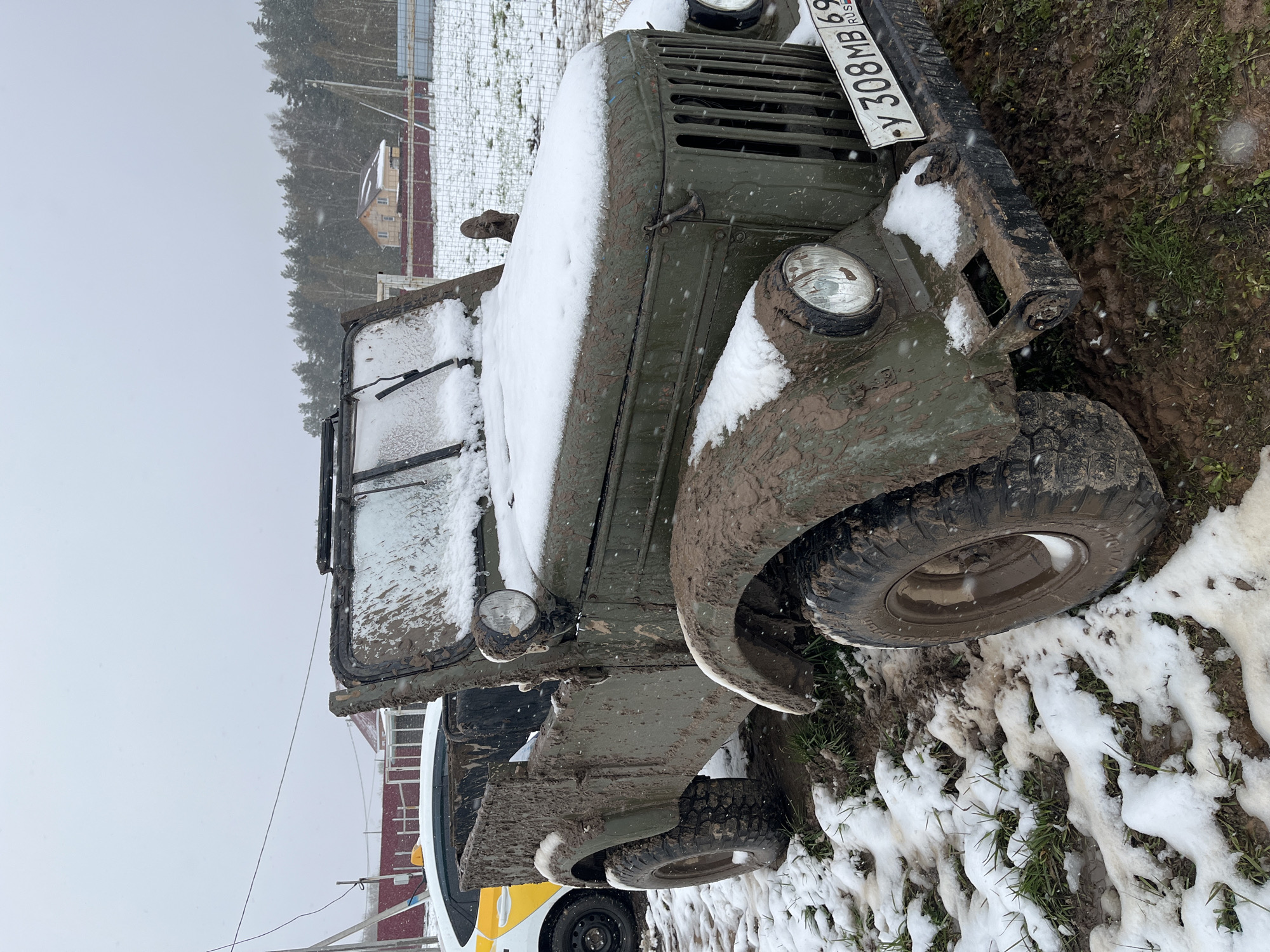 An olive-green, military-style utility vehicle, heavily caked in mud and partially covered in fresh snow, is the central subject of the image. The vehicle, likely a UAZ-469 or similar model, features large, knobby off-road tires and is equipped with a front license plate displaying "Y 308MB 69 RUSSIA." The "69" code indicates the Tver Oblast region of Russia, consistent with the provided location context of Razdolye.
The scene depicts a cold, overcast daytime in what appears to be a rural or semi-rural setting. Light snow is actively falling, visible as small specks, and has accumulated on the vehicle's hood, fenders, and windshield. The ground around the vehicle is a mix of deep mud, patches of green grass, and melting snow, with visible tire tracks in the slush. In the background, a line of coniferous trees stands against the gray sky. Faintly visible beyond the trees are a few buildings, possibly houses or structures, and parts of a fence. Another vehicle, white and yellow, is partially visible behind the green off-roader. The overall impression is one of rugged outdoor conditions in winter or late autumn.