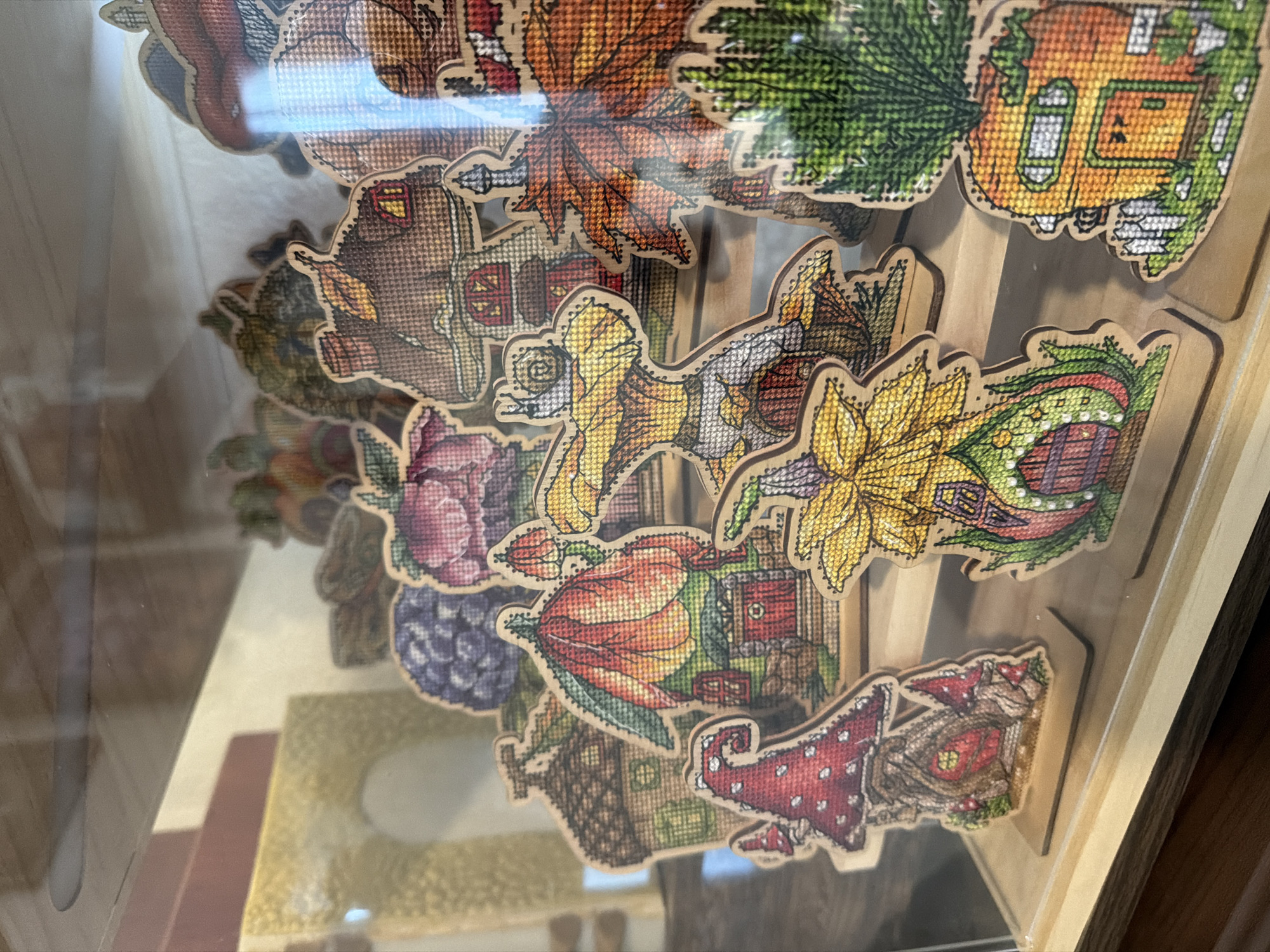 The image shows a display of miniature, cross-stitched houses shaped like various natural elements such as mushrooms, flowers, leaves, and fruits. These colorful, whimsical structures are arranged on wooden shelves within a clear display case. The houses are detailed with small doors, windows, and decorative elements, creating a fantasy village scene. The background is blurred but suggests an indoor setting with warm lighting. The image is taken from a slightly elevated, close-up perspective, focusing on the intricate details of the miniature village. There are no people visible in the image. The location of this display is in Aliso Viejo, United States of America.