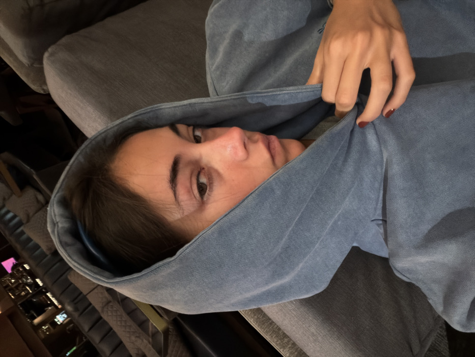 A young female is photographed in an indoor environment, identified as Mexico City, Mexico. She is seated or reclining on a gray upholstered surface, with her upper torso and face prominent in the frame. She wears a light blue, faded hooded sweatshirt with the hood drawn over her head. Her dark brown hair is visible under the hood, along with a light blue headband. Her right eye exhibits a visible swelling on the lower eyelid. Her right hand, adorned with dark red nail polish, is seen holding the fabric of her hoodie near her chest. The background is blurred, revealing multiple dark upholstered seating units, possibly sofas or benches with quilted detailing, in a dimly lit setting. The subject maintains a direct, neutral gaze towards the camera.