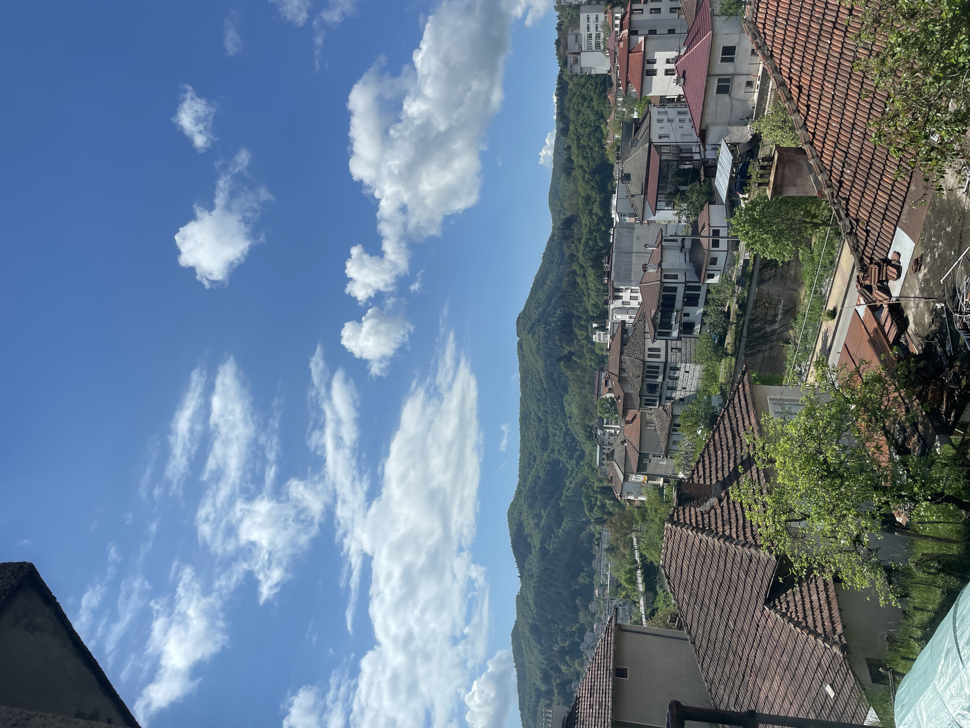The image shows a view of Zlatograd, Bulgaria. In the background, a forested mountain slope rises. In the midground, buildings and houses with red-tiled roofs are situated, interspersed with greenery. The foreground shows a red-tiled roof of a building. The sky is blue with white clouds.