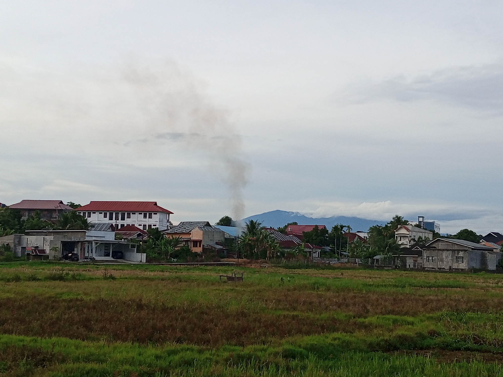 The image depicts a landscape scene in the city of Cotgapu, Indonesia, under an overcast sky. In the foreground, a field of green and brown vegetation stretches across the frame. Beyond the field, a cluster of buildings is visible, featuring houses with red and white roofs, some with multiple stories. One building is a multi-story white structure with many windows. Further in the background, a mountain range is visible, partially obscured by clouds. A plume of dark smoke rises into the sky from behind the buildings, suggesting an ongoing fire or industrial activity. The overall scene is a mix of residential structures, natural landscape, and a sign of human activity.