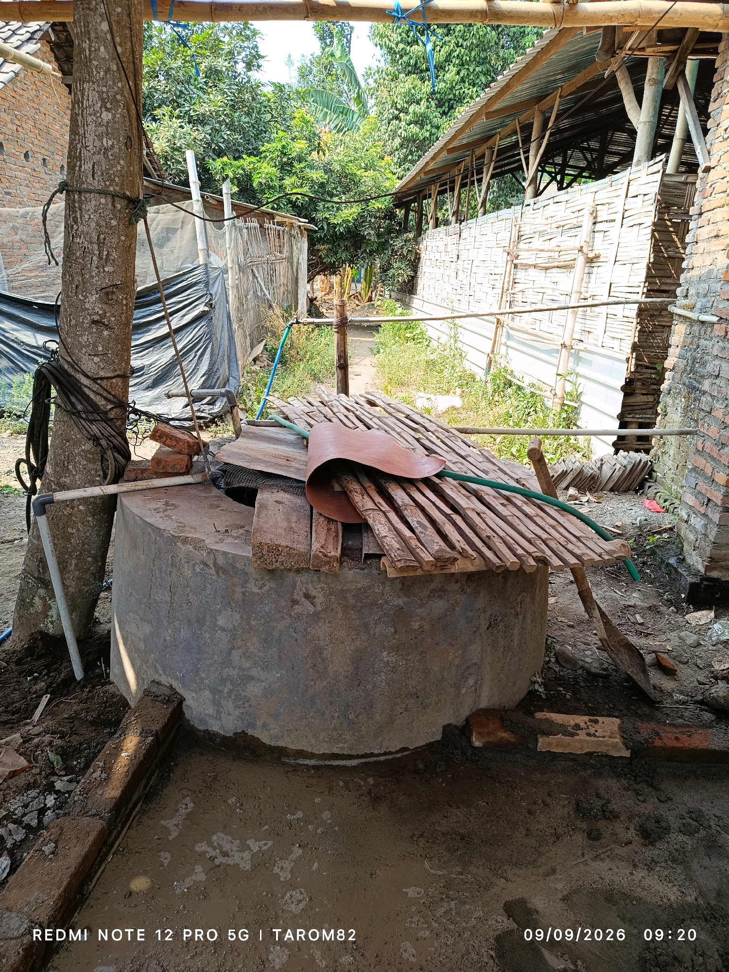 In Rejoagung, Indonesia, a well constructed from concrete stands in a rural setting. The well is covered with a wooden platform, suggesting it is either under construction or being maintained. A green hose runs over the wooden cover, possibly for filling or emptying the well. Several lengths of bamboo are also part of the covering. To the left of the well, a large tree trunk serves as a support for various cables and ropes, some of which are attached to a black tarpaulin structure behind it. The area around the well is rough, with loose dirt, bricks, and a shovel scattered about, indicating ongoing work. In the background, there are structures with tin roofs and walls made of woven bamboo, and lush green foliage suggesting a tropical environment. The sky is visible through gaps in the structures, appearing bright and clear. A brick wall is visible on the far right of the image. The bottom of the image displays a timestamp, reading "09/09/2026 09:20".