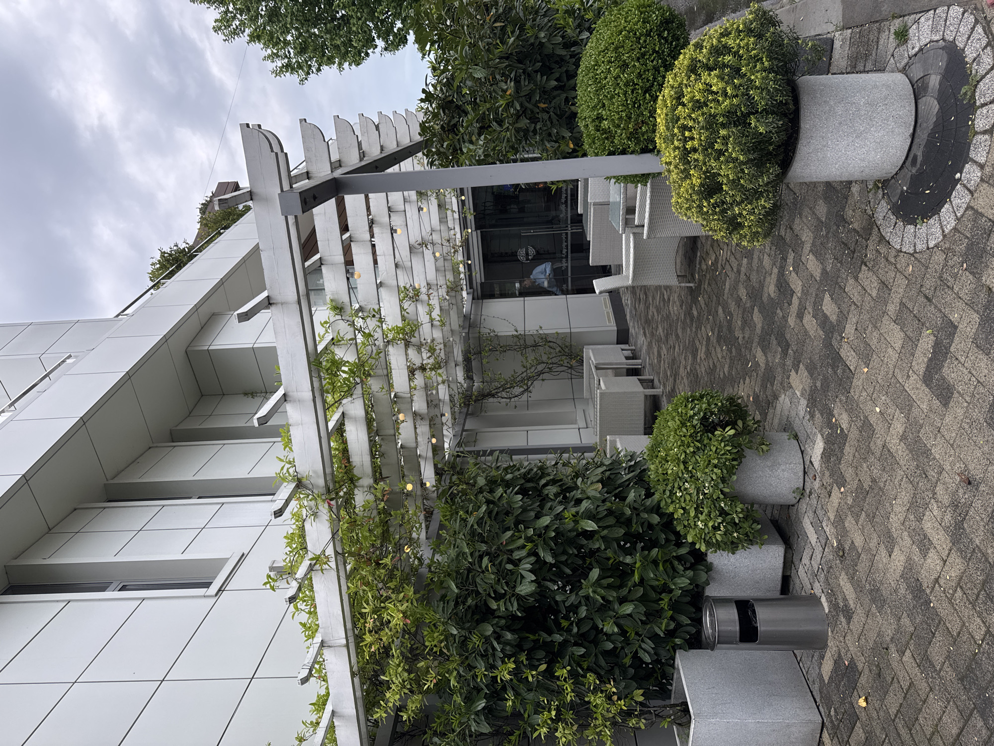 The image shows an outdoor seating area adjacent to a modern, white building. The seating area is covered by a white pergola structure with vines growing on it, adorned with small lights. The area is furnished with white wicker chairs and tables. Potted plants and hedges provide greenery. A cylindrical gray trash receptacle stands near one of the planters. The pavement is patterned in gray bricks. The sky is overcast. Reflection in the glass door suggest the presence of a person inside. Based on visual cues, location information is impossible to determine from the provided image. No specific indicators point to Sochi, Russia.