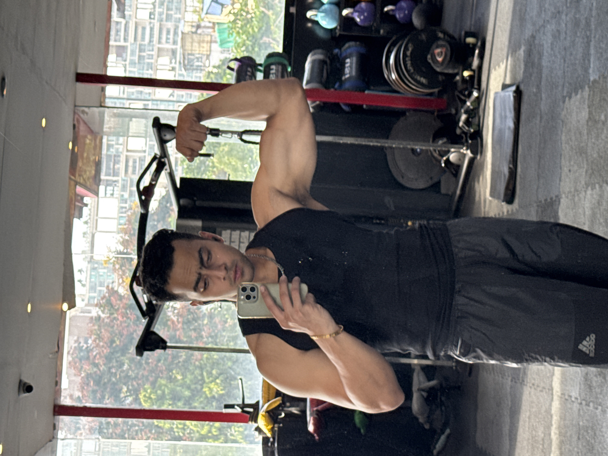 In a gym in Kolkata, India, a man is taking a selfie. He is wearing a black tank top and dark gray shorts. His right arm is flexed, showing off his bicep. He is holding a smartphone in his left hand, which is pointed towards the mirror to capture his reflection. The gym equipment, including weight plates and kettlebells, is visible in the background. A security camera is mounted on the ceiling. The man appears to be in his 20s or 30s. The gym has large windows that offer a view of buildings and trees outside. The floor is covered with interlocking mats.
