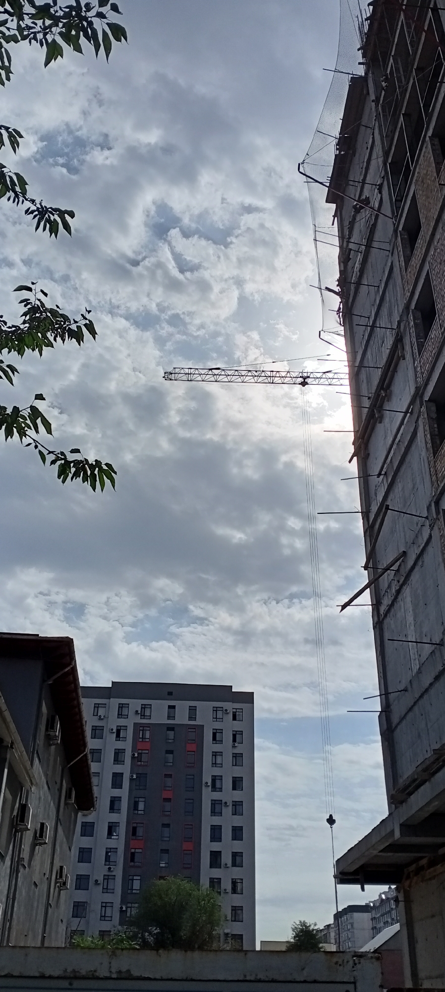 In Bishkek, Kyrgyzstan, an outdoor, low-angle shot captures a construction scene dominated by a sky filled with dense, gray clouds. To the right, a multi-story building under construction is prominently featured, its concrete frame and brickwork visible. Safety netting is draped over the upper floors. Extending horizontally across the sky, a construction crane is suspended, with its boom silhouetted against the bright, hazy sun that peeks through the clouds. To the left of the crane, a branch with green leaves intrudes into the frame from the top-left corner. In the mid-ground, a completed, modern residential building with a gray and red facade stands with numerous windows. In the foreground, a portion of another building with a darker exterior and a red-tiled roof is visible on the far left. A low wall or barrier runs along the bottom of the image. The overall impression is one of ongoing urban development under a dramatic sky.