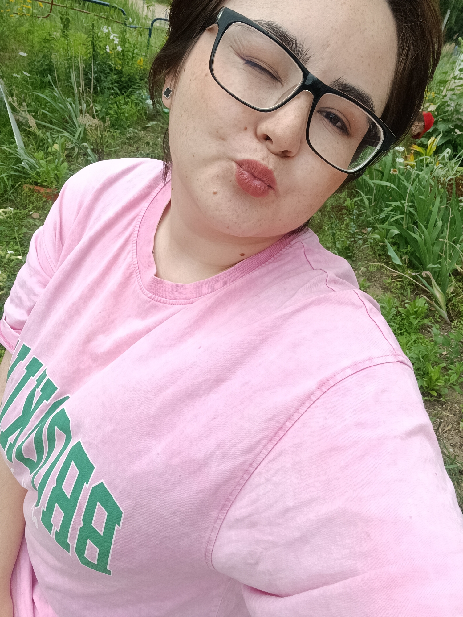 A young woman with light skin and dark hair is taking a selfie outdoors in front of a garden with lush green foliage. She is wearing a pink t-shirt with a green, collegiate-style text graphic on it. The text appears to read "BROOKLYN". She has on black-rimmed glasses and is winking and puckering her lips as if blowing a kiss. She has a smattering of freckles across her cheeks and nose. The setting appears to be a private garden or yard, with various plants and some gardening tools visible in the background. The lighting suggests it is daytime, possibly a sunny day. The overall mood conveyed is playful and cheerful.
