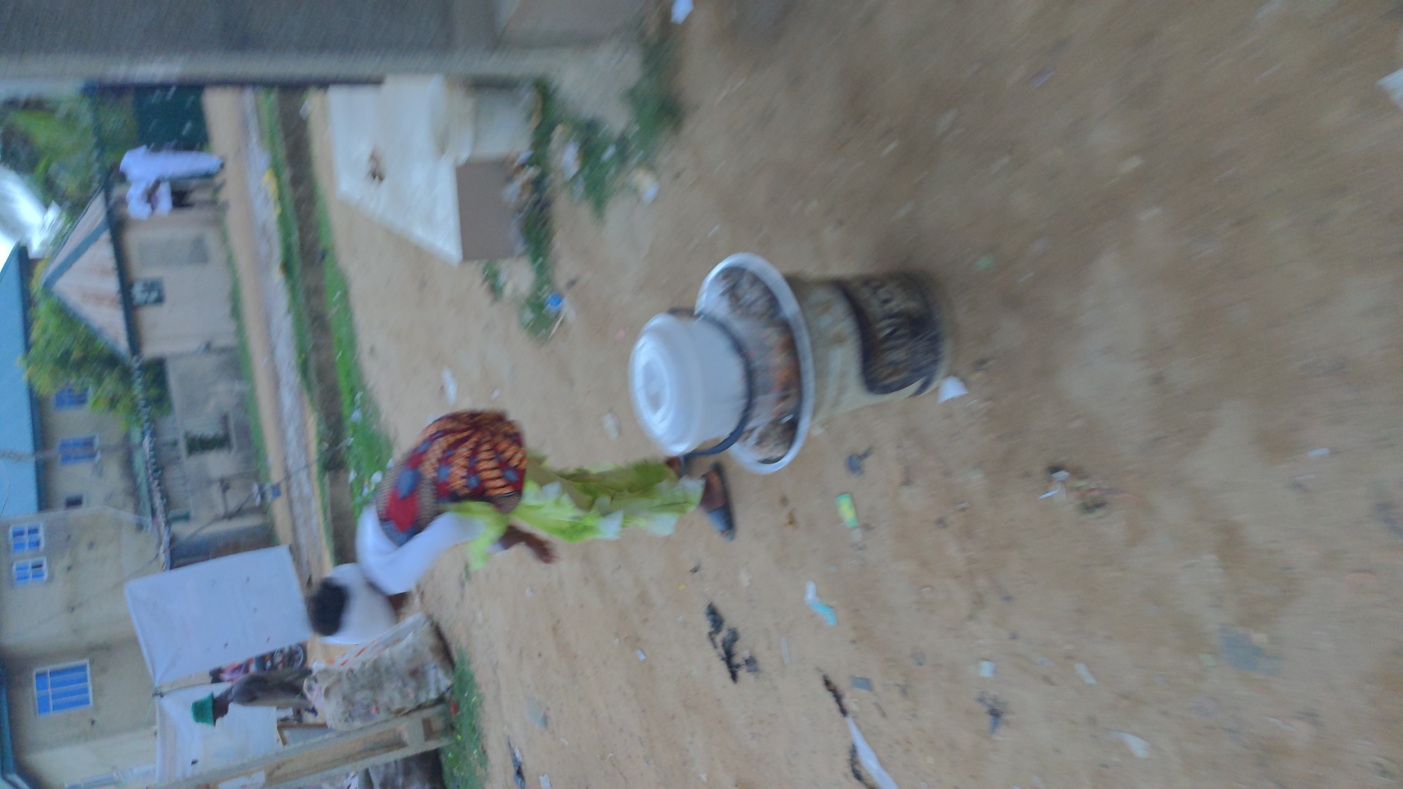 The scene depicts a woman carrying a load on her head, walking on a sandy surface in Ado, Nigeria. She is wearing a patterned headscarf, a white top, and a green wrap around her waist. The load on her head appears to be a layered container, potentially holding food or goods. The background shows buildings of varying heights and construction styles, characteristic of urban areas in Nigeria. Debris is scattered on the ground. Another person can be partially seen in the lower left of the frame.
