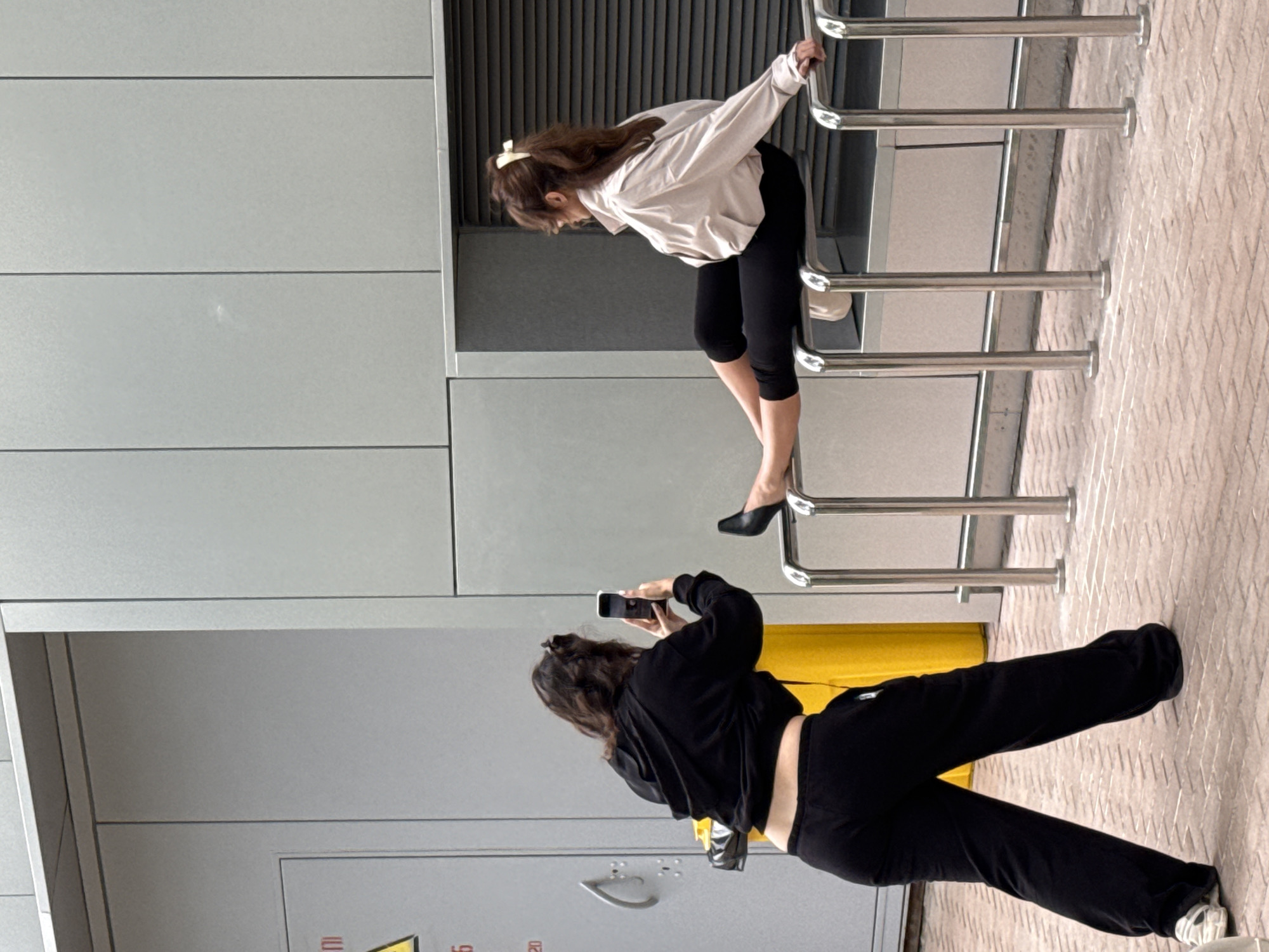 The image shows two young women in an outdoor setting, likely in Moscow, Russia. The scene appears to be near a modern building with light grey panels and a metallic grate in the background. The ground is paved with diamond-patterned tiles.
In the foreground, a woman wearing a black hoodie and black pants is captured from the back, holding a smartphone and taking a picture of the other woman. Her body is angled to the side, suggesting she is focused on her photography.
In the background, a second woman, with long brown hair tied with a light-colored bow, is leaning forward over a series of parallel metal railings. She is wearing a light-colored, possibly beige, long-sleeved top and black cropped pants. Her black high-heeled mules are visible. Her posture suggests she is posing or interacting with the railings.
The lighting indicates it is daytime, and the weather appears to be fair. There are no prominent emotional expressions visible due to the framing and focus of the image. Visible text is minimal, with what appears to be partial Cyrillic lettering on a yellow object in the background. The overall scene suggests a casual outing or photo session.