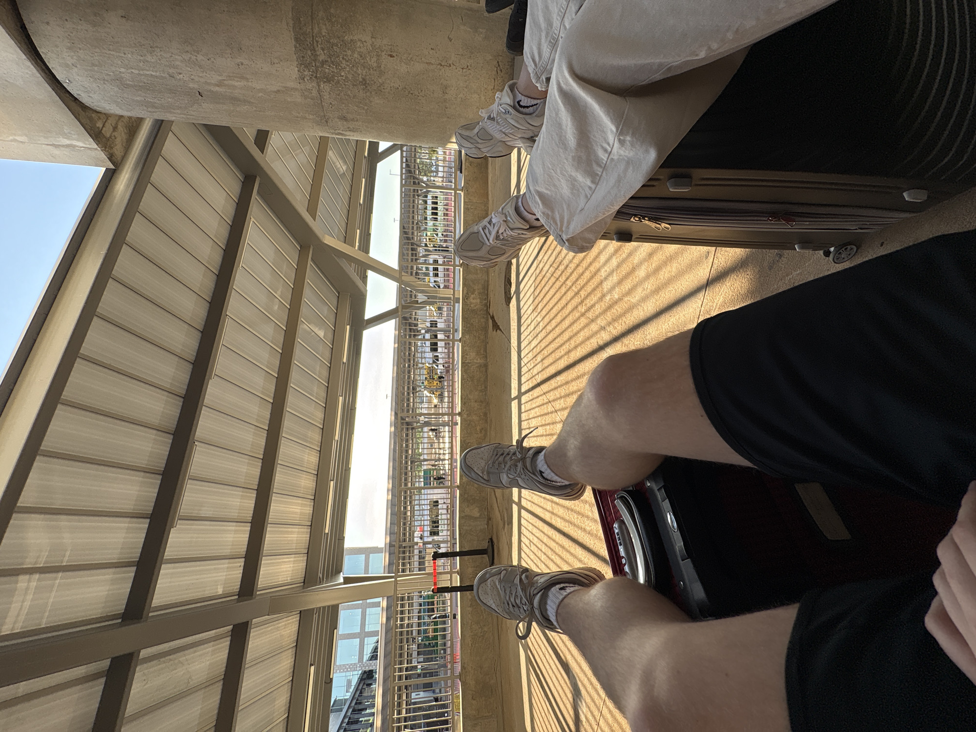 The image, taken from a low angle, depicts a partially enclosed outdoor space with a corrugated metal awning above. Two individuals are visible from the knees down, sitting on luggage. One person is wearing light-colored pants and sneakers, seated on a tan suitcase with wheels. The other person is wearing black shorts and sneakers, situated on a dark suitcase. The scene is illuminated by sunlight casting shadows across the ground. In the background, a cityscape is visible through the openings of the structure, potentially identifying the location as an outdoor area in the city of College Park, United States of America.