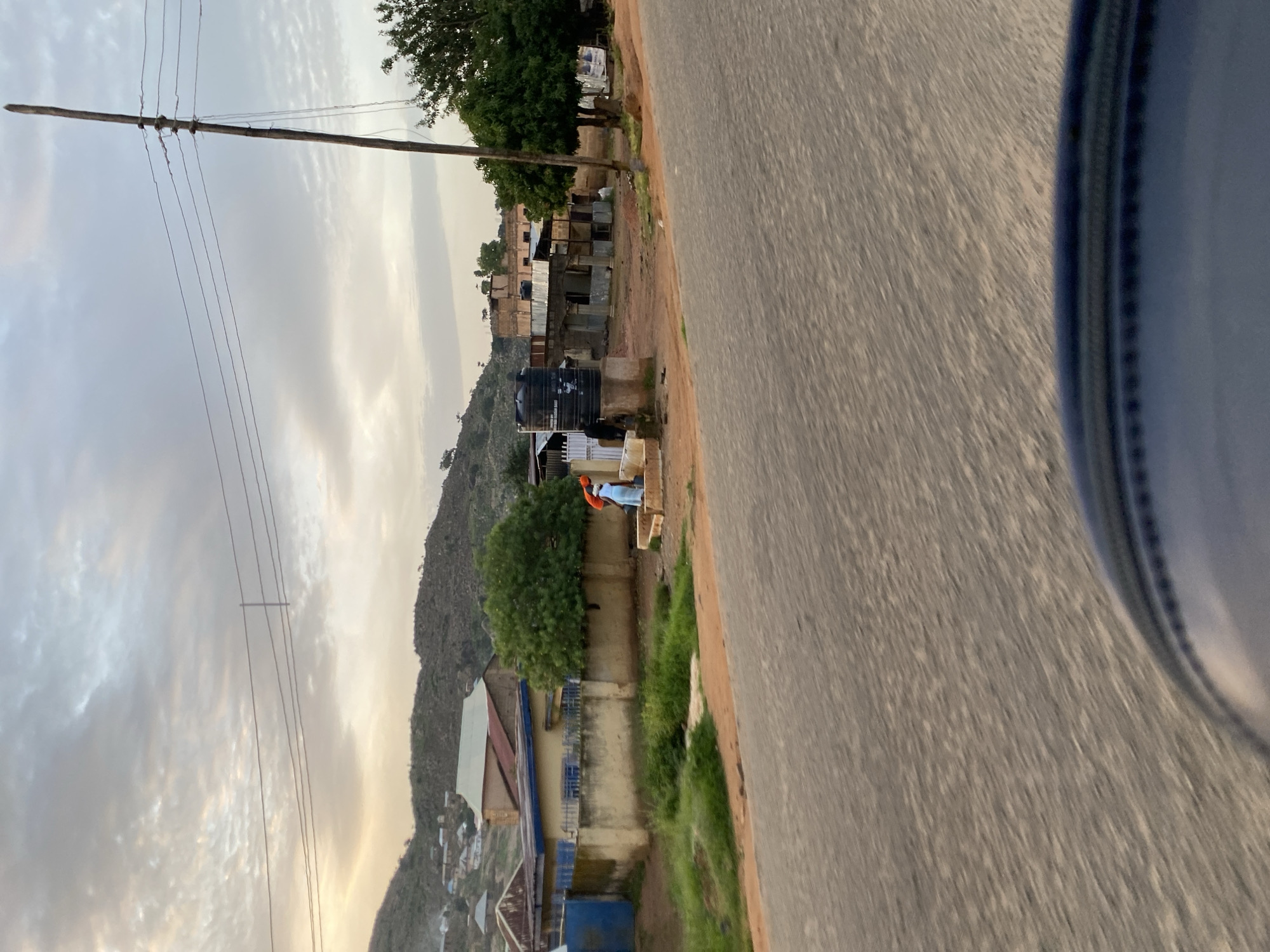 The image captures a view from a moving vehicle in Jos, Nigeria, looking towards a residential area nestled against a hilly backdrop. The foreground is dominated by a blurred asphalt road, with the edge of what appears to be a vehicle's dashboard or seat visible in the bottom right.
In the mid-ground, a cluster of buildings lines a dirt road. These structures vary in architectural style, from simple, single-story homes with corrugated metal roofs to more complex, multi-story buildings. A prominent element is a large black water tank situated on a raised platform. Two individuals are visible near this tank; one, wearing an orange garment, appears to be interacting with a second person in a light blue garment. They are positioned on a small set of steps.
Further back, the landscape rises into rolling hills, sparsely populated with smaller dwellings and scattered trees. A tall, wooden utility pole with electrical wires crisscrossing overhead stands prominently on the right side of the frame, indicating the presence of infrastructure. The sky is overcast with textured clouds, suggesting either early morning or late afternoon light. The overall impression is of a developing urban periphery with elements of rural life integrated into the landscape.