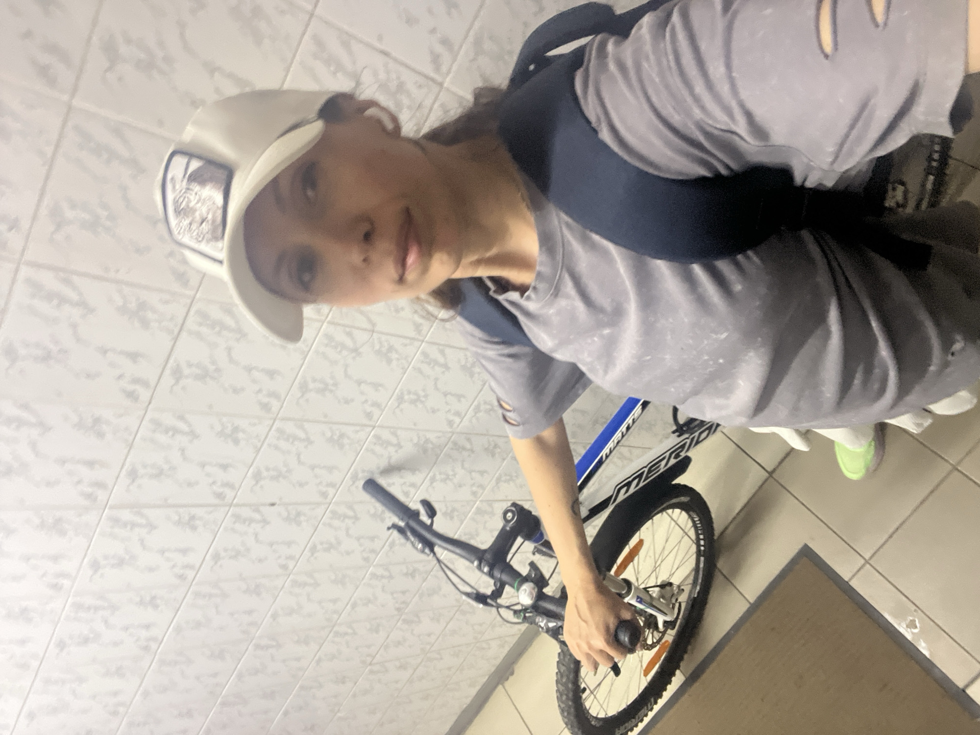 A woman with a white baseball cap and gray t-shirt stands with a black backpack, next to a Merida mountain bike in an indoor space with tiled walls and floors. The location is Moscow, Russia.