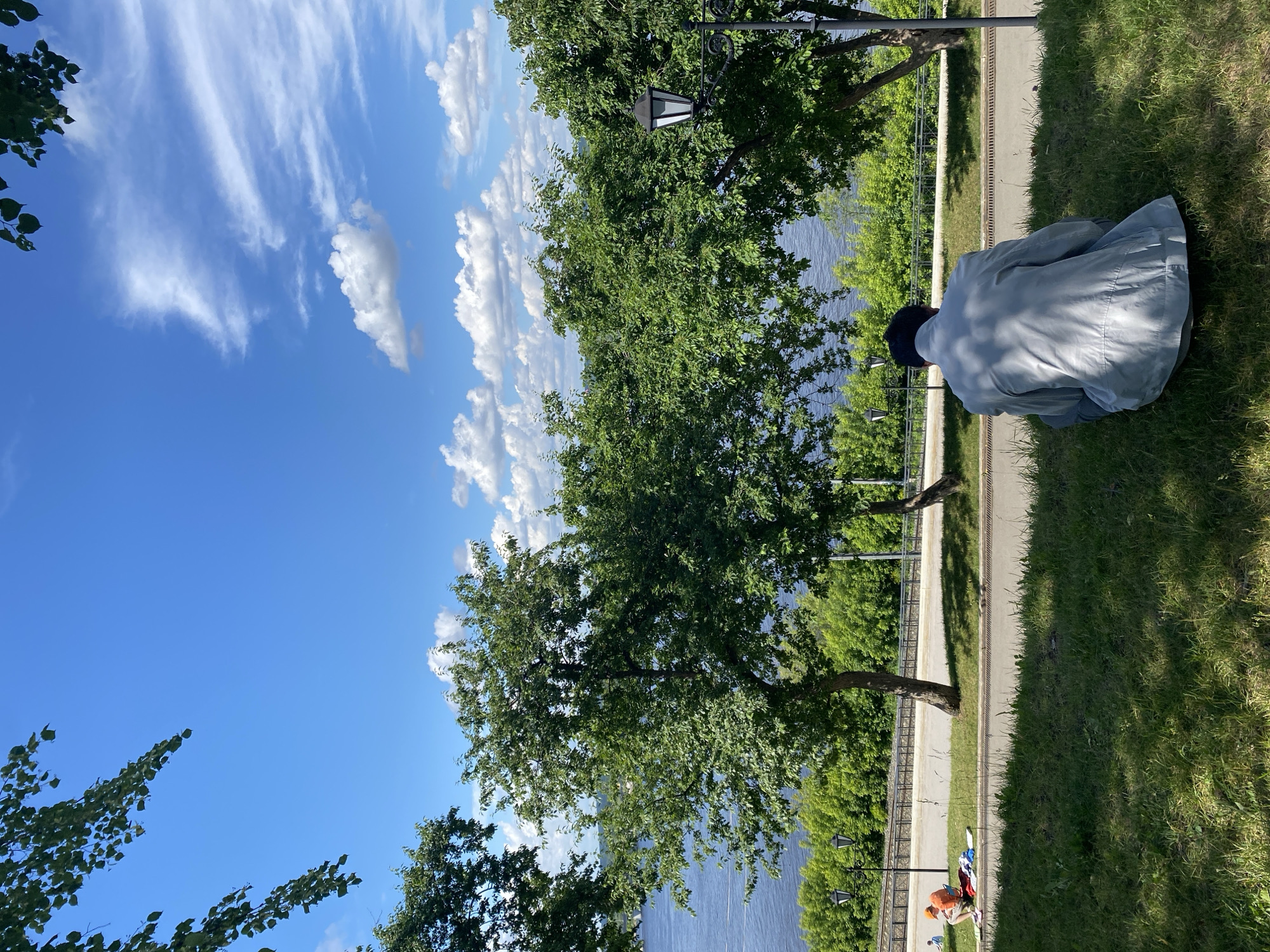The image depicts a person sitting on the grass in the foreground, facing away from the camera and towards a river and a treeline. The person is wearing a light-colored jacket and dark pants. In the middle ground, there is a paved pathway with trees lining the edge and a river beyond. Further in the background, there are more trees and a bright blue sky with scattered clouds. To the left of the image, there are two people sitting on the grass near the pathway. This scene is consistent with the area along the Kama River embankment in Perm, Russia.