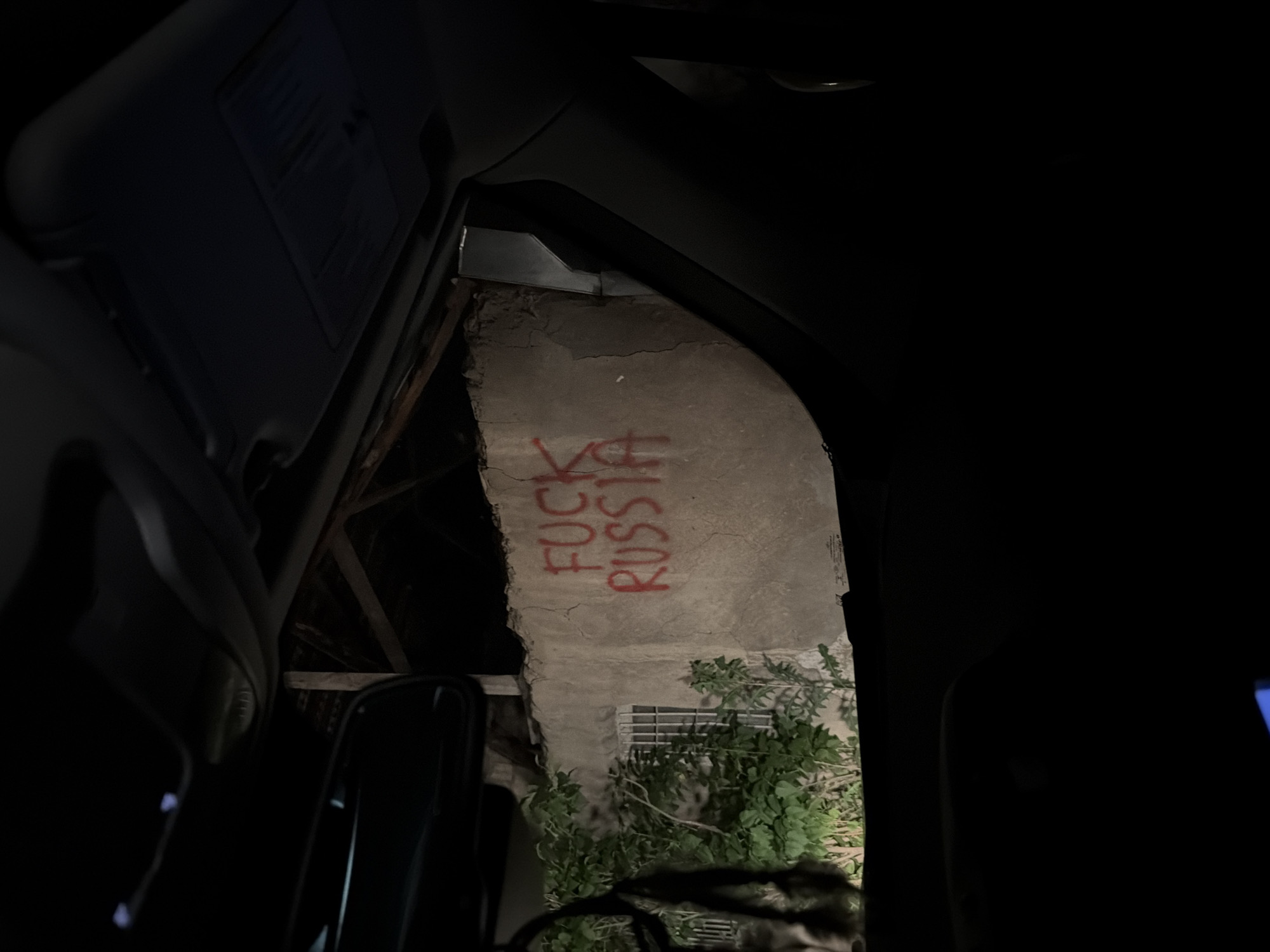The image, taken at night from inside a vehicle, shows a view of a building exterior with the phrase "FUCK RUSSIA" spray-painted in red. The building is made of concrete or stone, with visible cracks and aged appearance. Part of a barred window is visible beneath the graffiti. Below the window, there are leafy green plants. The vehicle interior includes the sun visor and part of the driver's seat. Based on the visible graffiti, the image is likely taken in Tbilisi, Georgia.