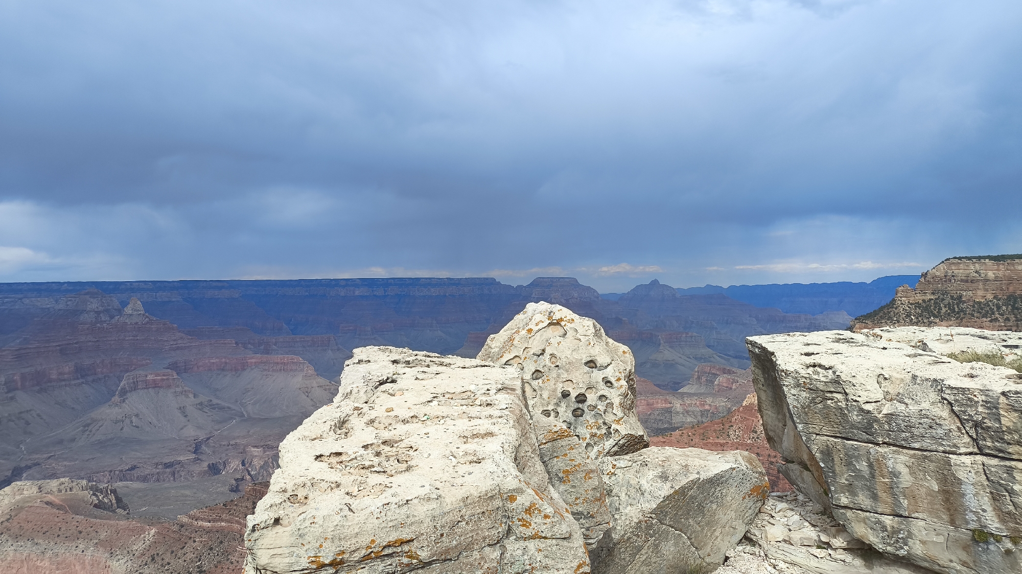 A wide-angle, eye-level shot captures a dramatic vista of the Grand Canyon under a cloudy, overcast sky. In the foreground, two large, weathered white rocks dominate the scene, their surfaces textured and showing hints of orange lichen. The rock on the right is larger and more angular, with visible cracks and striations. The rock on the left is flatter and more rounded.
Beyond the foreground rocks, the vast expanse of the Grand Canyon unfolds. Layers of reddish-brown and purple rock formations stretch out into the distance, sculpted by erosion into mesas and cliffs. The distant canyon walls fade into a hazy blue, indicating depth and scale. The sky above is a heavy blanket of dark gray clouds, with lighter patches suggesting breaks in the overcast. There are no people or animals visible in the image. The overall impression is one of natural grandeur and the powerful forces of geological time.