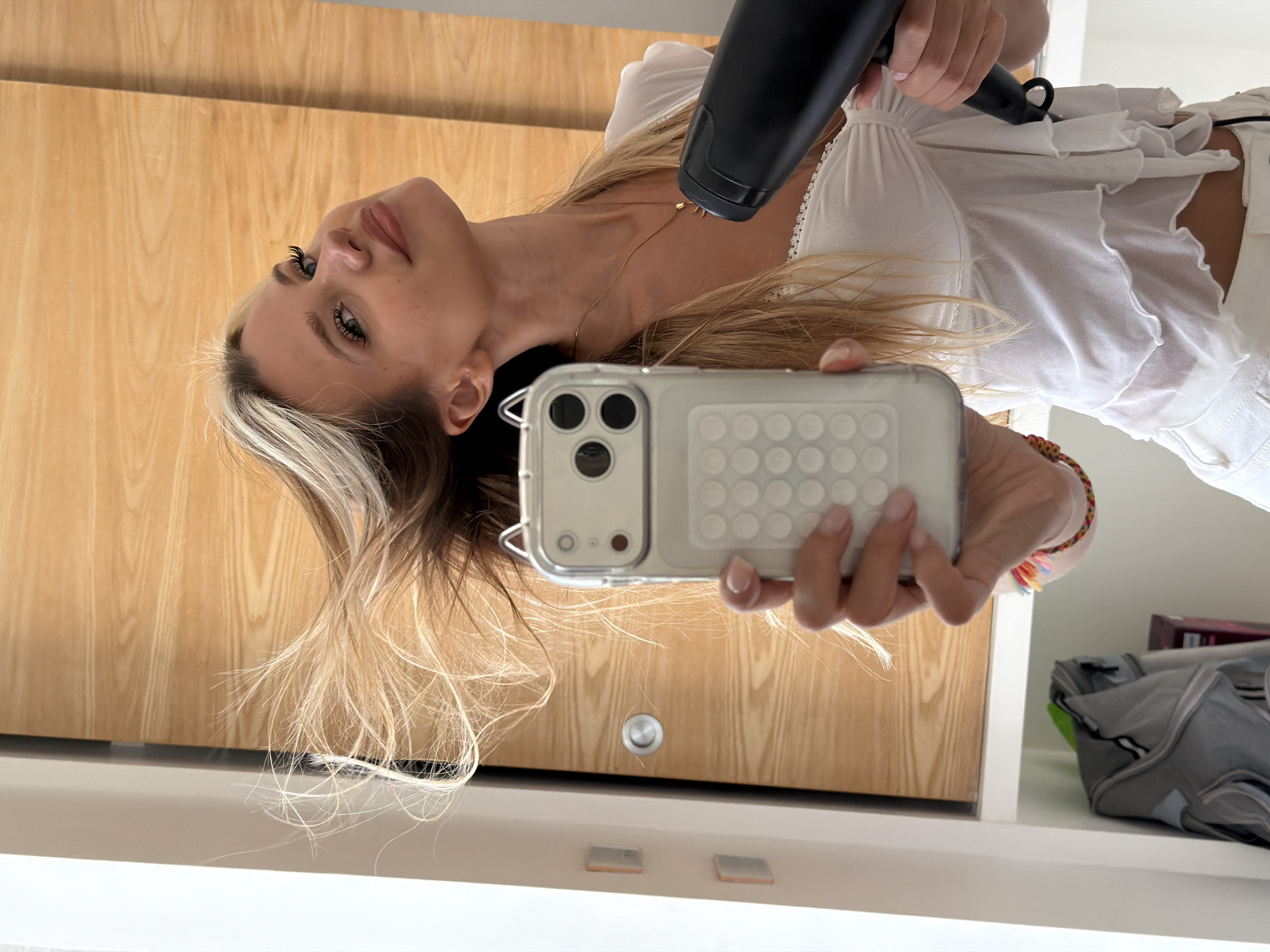 A young woman with blonde hair is seen from the chest up, holding a smartphone in her left hand and a black hairdryer in her right hand. Her hair is being blown upwards by the hairdryer, which is pointed towards her head from the right side of the frame. She is wearing a white, sheer top with ruffles. She is looking at the camera and smiling. The background consists of a wooden door and a white wall. There is a grey backpack hanging on the wall to the left. The scene is indoors, likely a bathroom or bedroom, with bright lighting. The location in the city of Thep Krasatti, Thailand is not discernible from the image itself.