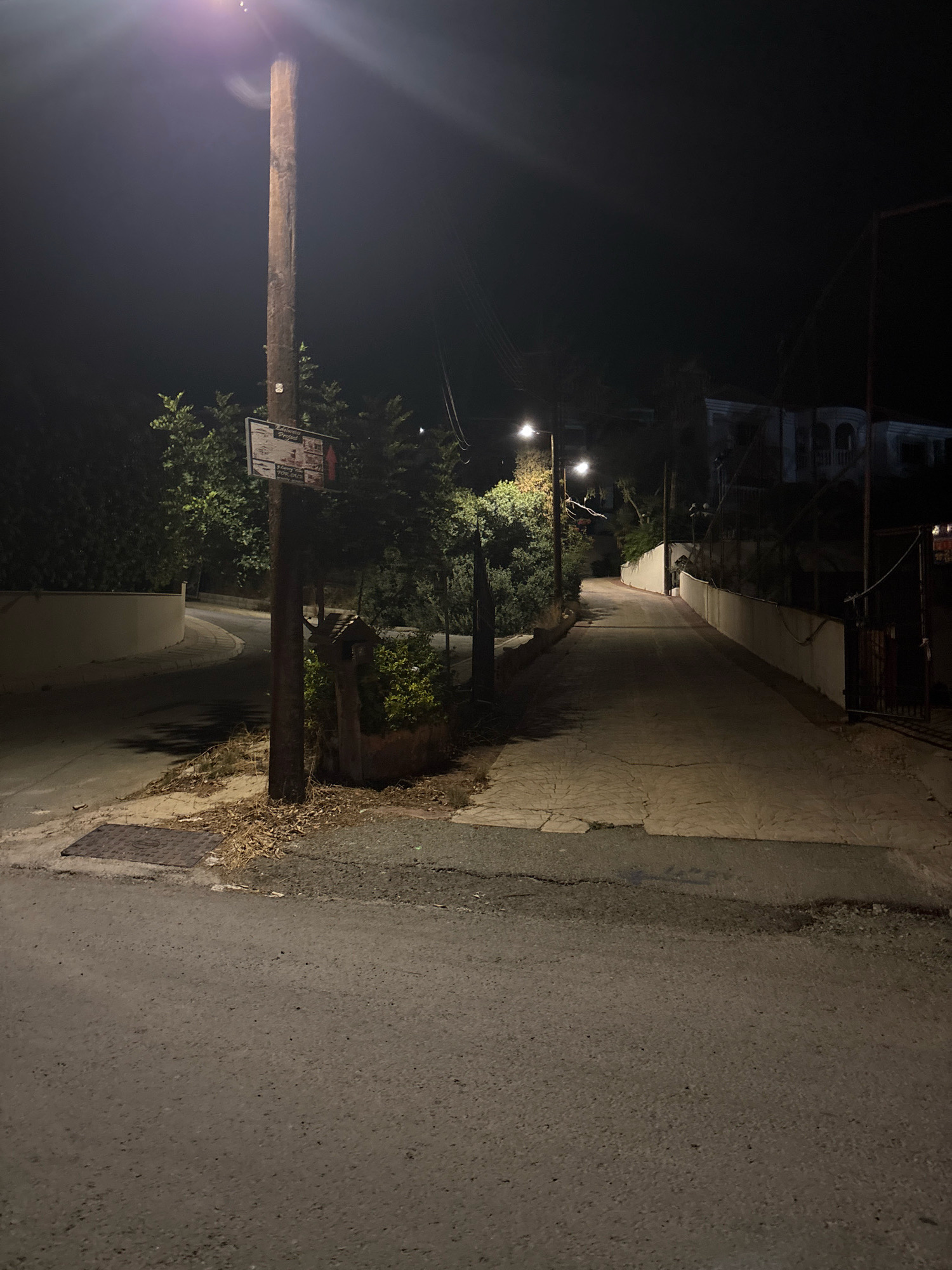 A low-angle night shot captures a dimly lit, paved road in Agios Tychona Community, Cyprus. The road curves to the left in the foreground and continues straight into the background, lined by low white walls and dense green foliage on either side. A wooden utility pole stands prominently on the left, illuminated by a bright overhead streetlamp that casts a purple glow and lens flare across the upper portion of the frame. Below the light, a sign is attached to the pole. Two more streetlights are visible further down the road, casting pools of yellow light. A rusty metal gate and fence are visible on the right side of the road in the background. The ground is a mix of asphalt and textured paving stones, with dry leaves and dirt scattered near the utility pole and a metal grate in the road. The overall scene depicts a quiet, residential street at night.