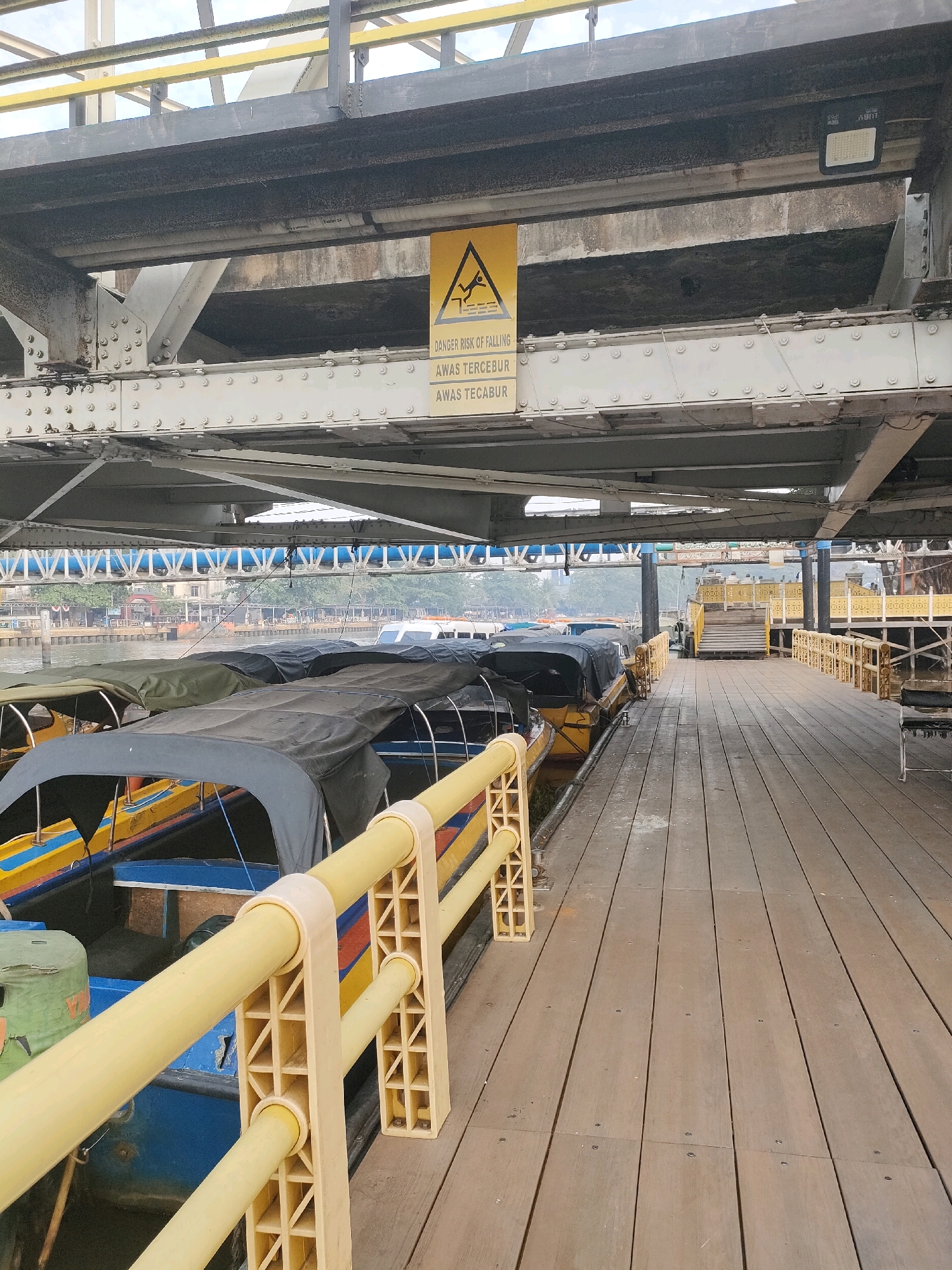 The image captures a view from a wooden pier in Banjarmasin, Indonesia, likely a river port or a docking area for boats. In the foreground, several colorful boats, covered with dark tarps, are moored along the pier. A bright yellow railing, made of plastic material with a lattice design, runs along the edge of the pier, separating it from the boats. The pier itself is constructed of weathered wooden planks, showing signs of age and wear.
Above the pier, a large metal structure, likely a bridge or overpass, is visible. Hanging from this structure is a yellow warning sign with a black pictogram depicting a person falling. Below the pictogram, text reads "DANGER RISK OF FALLING" and in a different language "AWAS TERCEBUR" and "AWAS TECABUR", indicating a hazard of falling into the water.
In the background, across the river, a city skyline with buildings and trees is faintly visible through a light haze. Another pier or walkway with yellow railings and stairs is seen further down the riverbank. The overall scene suggests a busy, functional area for river transportation in the city. No people are visible in the image.