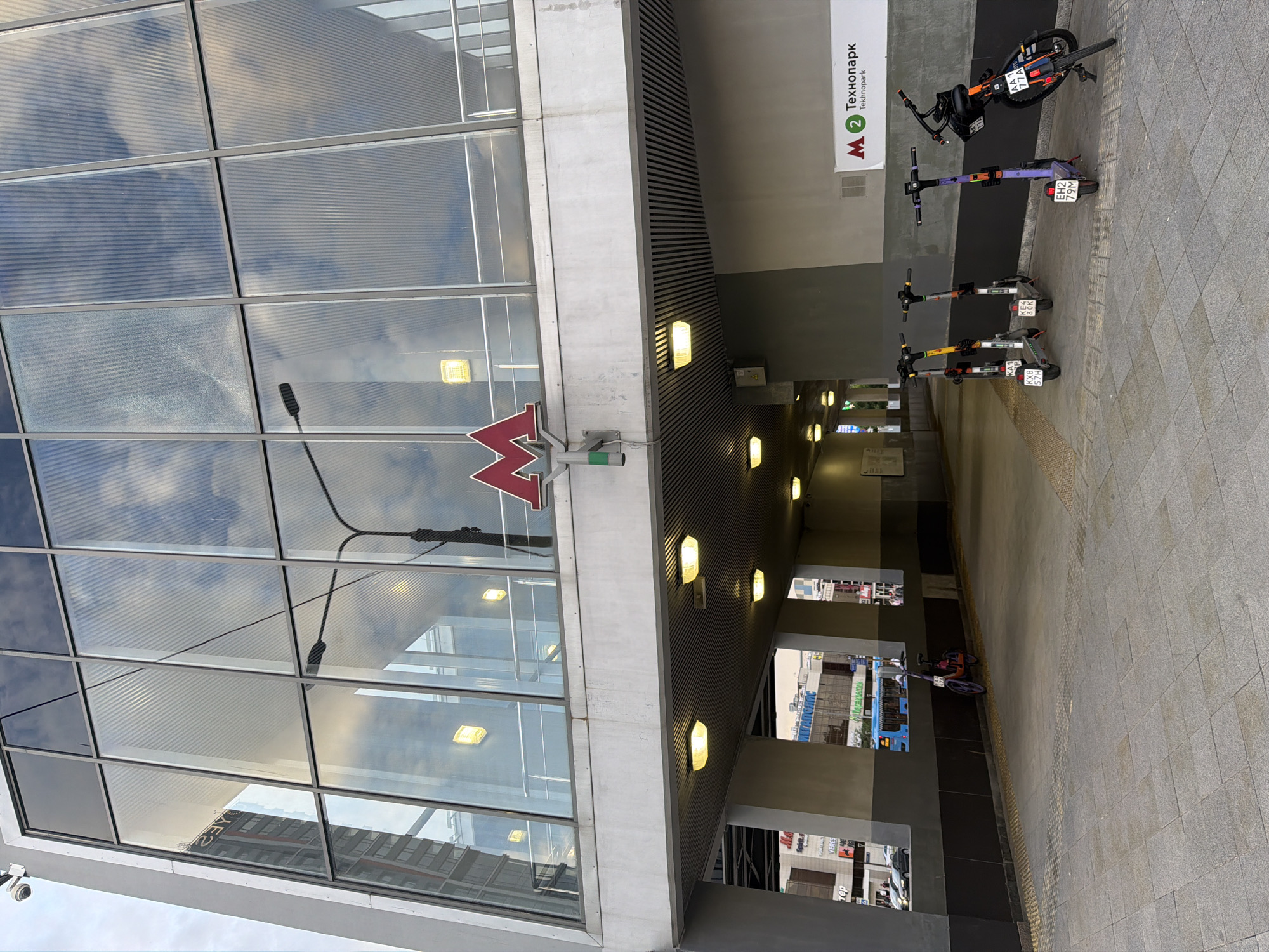 The image shows an outdoor view of the entrance to a metro station in a city. A large building with a modern facade of glass windows dominates the upper portion of the frame, reflecting the sky. Below the glass facade is a concrete structure with a covered walkway. Several electric scooters are parked in a row under the walkway. In the background, through an opening in the structure, a blue bus and other buildings are visible. A sign with the Moscow Metro logo and the station name "Технопарк" (Tekhnopark) is mounted on the wall. The ground is paved with grey tiles.
