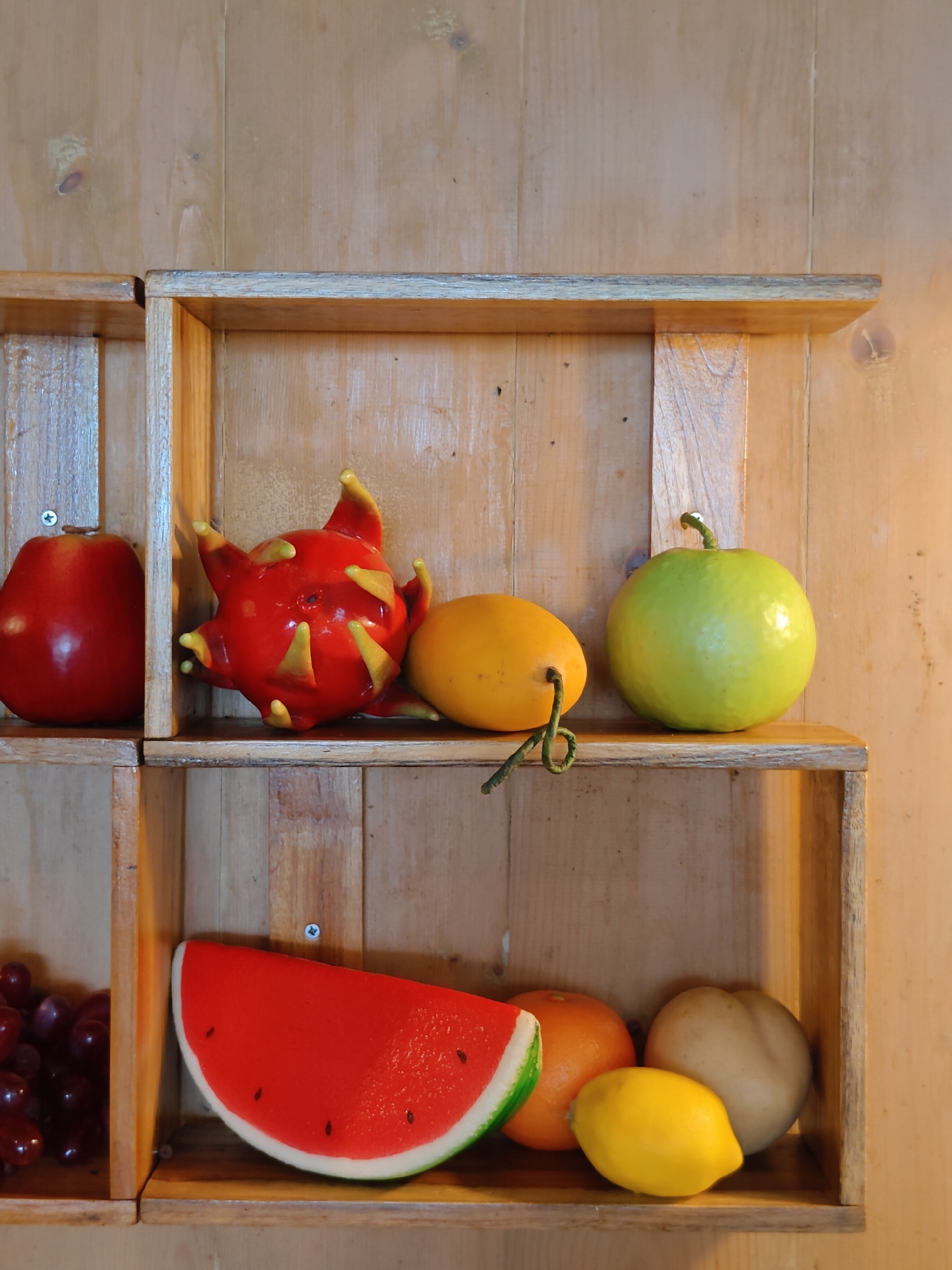 The image depicts a collection of artificial fruits displayed in wooden shelves against a wooden plank wall. The fruits include a red apple, a red dragon fruit, a yellow mango, a green guava, a slice of watermelon, a tangerine, a lemon, and a pear. The scene is illuminated by warm light, likely artificial, creating soft shadows. The wooden shelves are arranged in a two-by-two grid. The overall setting appears to be indoors. The location in Pattaya City, Thailand, is not discernable from the visual information provided in the image.