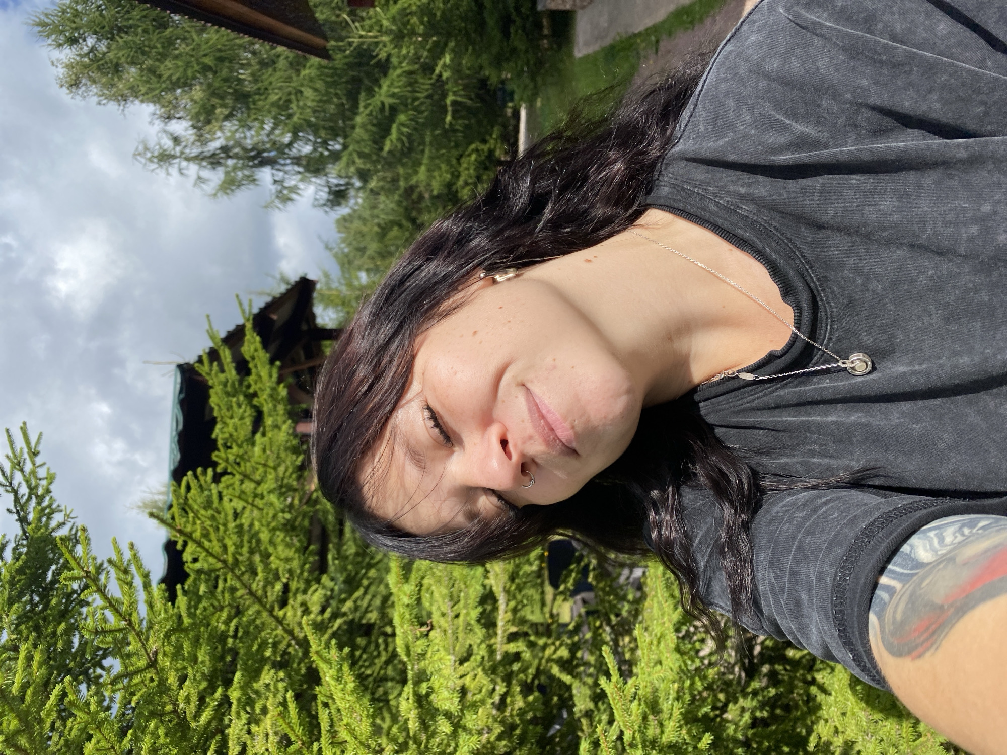A young woman with dark hair and closed eyes smiles in a selfie. She is wearing a black t-shirt and a silver necklace. Behind her, lush green trees and a cloudy sky are visible. In the background, wooden structures with green roofs suggest a rural or natural setting. The location is identified as Krasnoflotskoe, Russia.