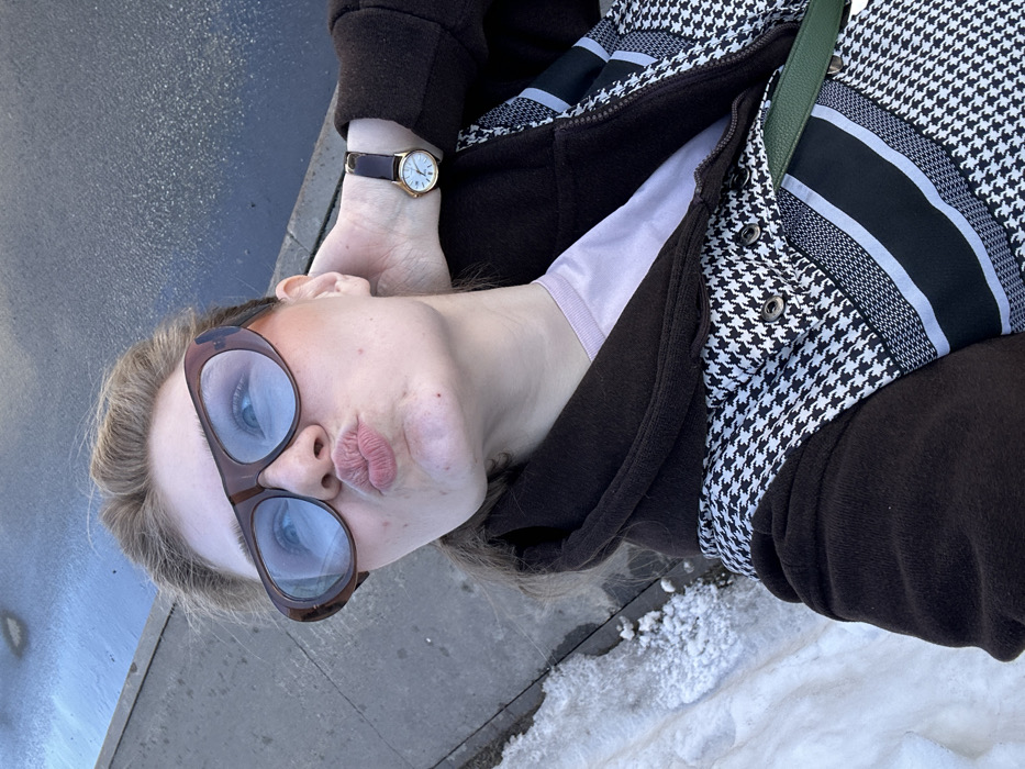 A female individual is depicted in an upward-angled close-up selfie, situated outdoors in Новое Шигалеево, Russia. She wears large, brown-framed sunglasses with blue-tinted lenses and has her lips puckered. Her right hand is positioned at the back of her neck. On her left wrist, she wears a gold-toned watch with a dark brown strap and a white face featuring Roman numerals. She is dressed in a light pink top under a jacket with dark brown sleeves and hood, and a torso section displaying a black and white houndstooth pattern accented by horizontal black and white striped details. Metallic snap buttons are visible on the jacket's front. A dark green strap is visible across her chest. The background shows a dark, reflective paved surface on the right and a patch of dirty snow or ice on the ground to the left, indicating a cold environment.