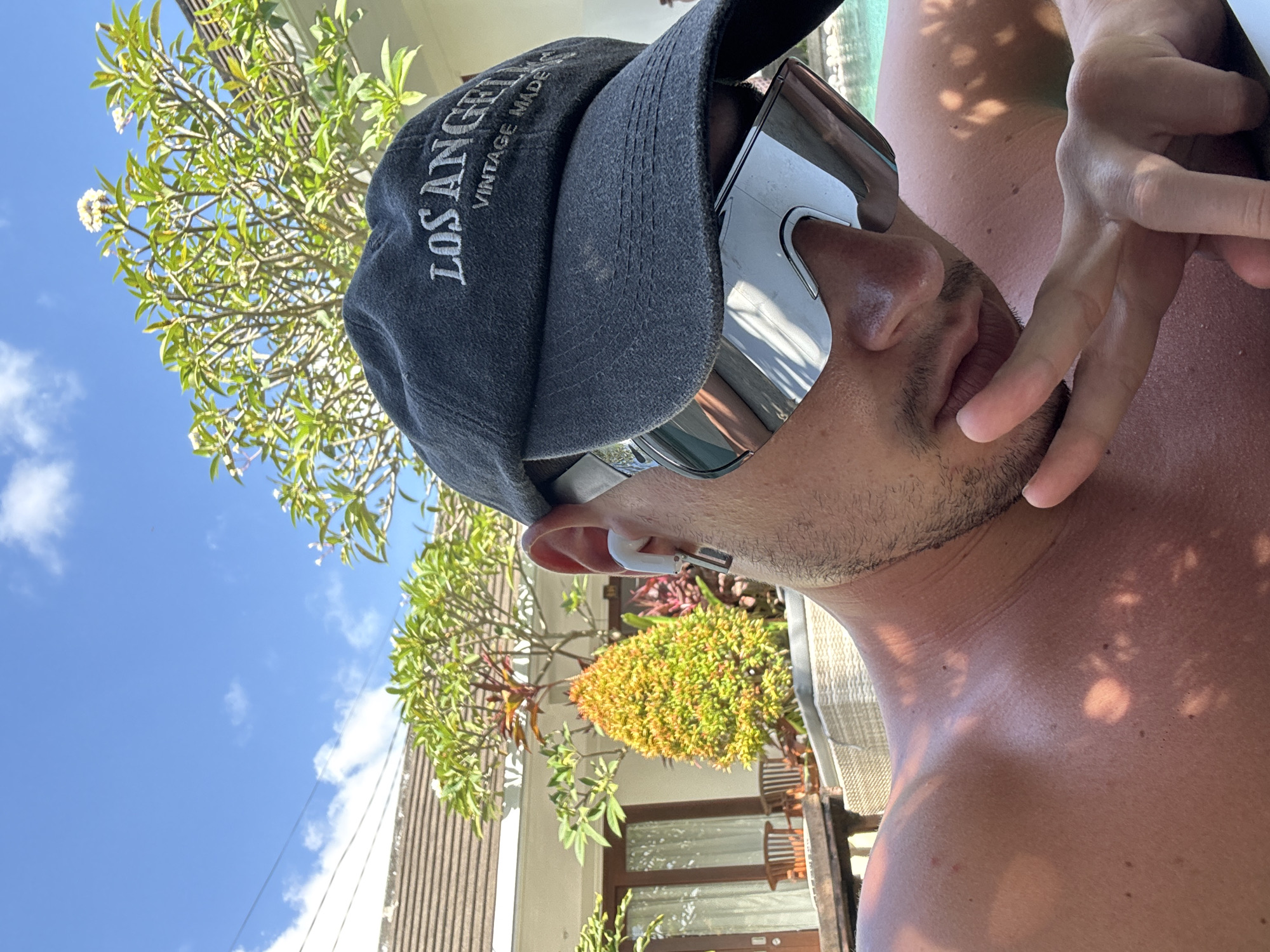 A man wearing sunglasses and a cap, with the words "LOS ANGELES VINTAGE MADE" on it, is looking at the camera. He is also wearing an earpiece. The setting appears to be outside, with foliage visible in the background, suggesting a garden or outdoor area in North Kuta, Indonesia.