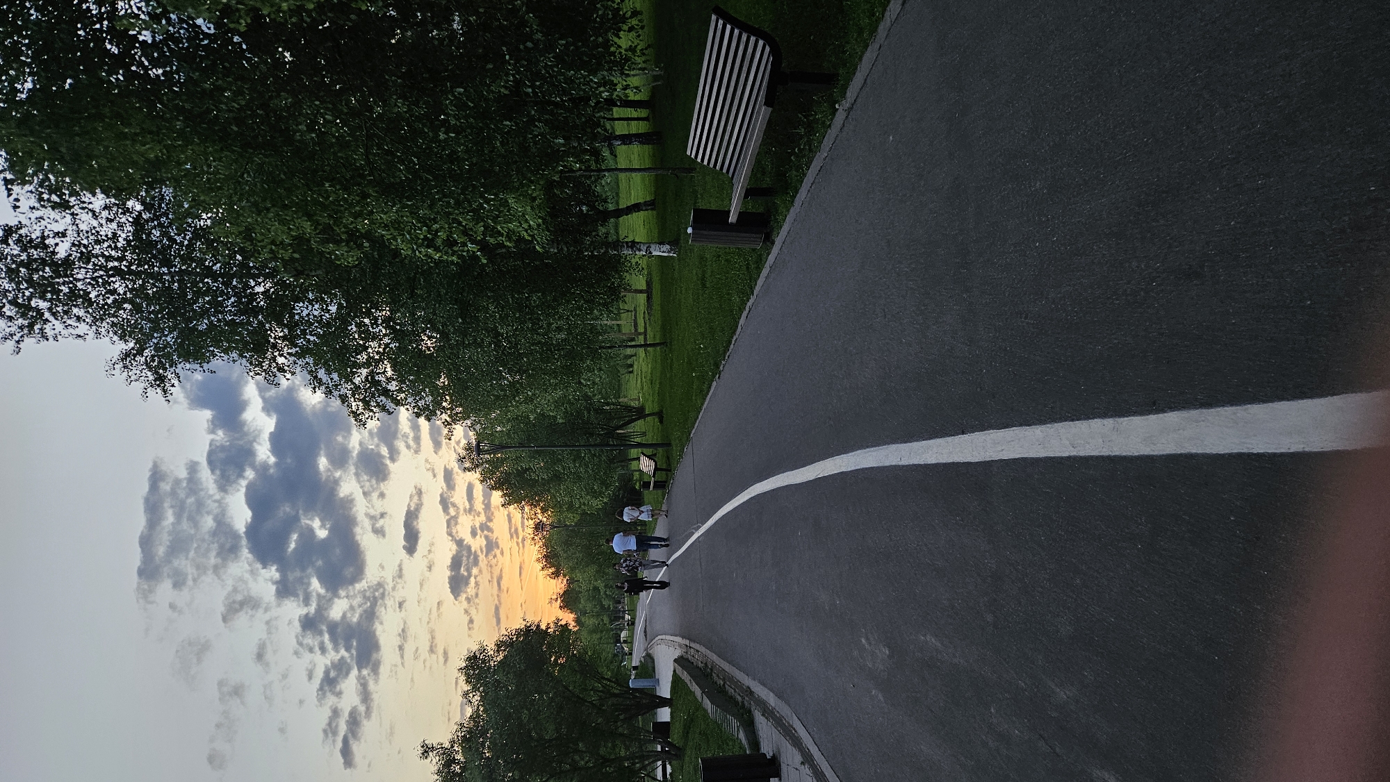 A dusk or dawn scene shows a curving asphalt pathway marked with a white line, flanked by grass and trees on one side and a bench on the other. Several people are seen in the distance on the pathway. The sky above features clouds tinged with orange and blue.