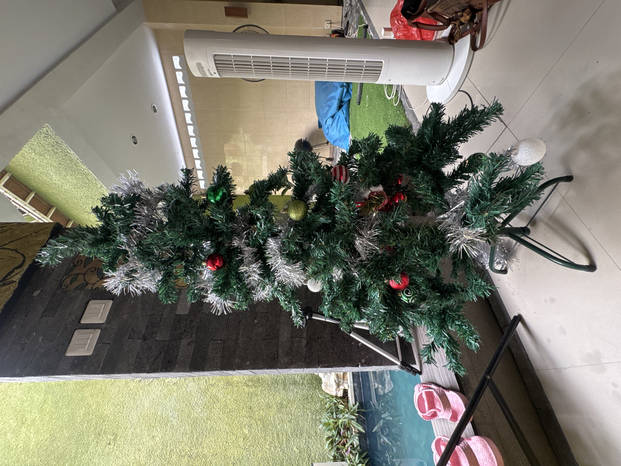 The image shows a Christmas tree indoors. It is decorated with silver tinsel and various colored ornaments, including red, green, and white. Near the tree is a white air purifier. In the background, there is what appears to be a small pool or water feature with pink sandals next to it. The location appears to be Kuta Selatan, Indonesia.