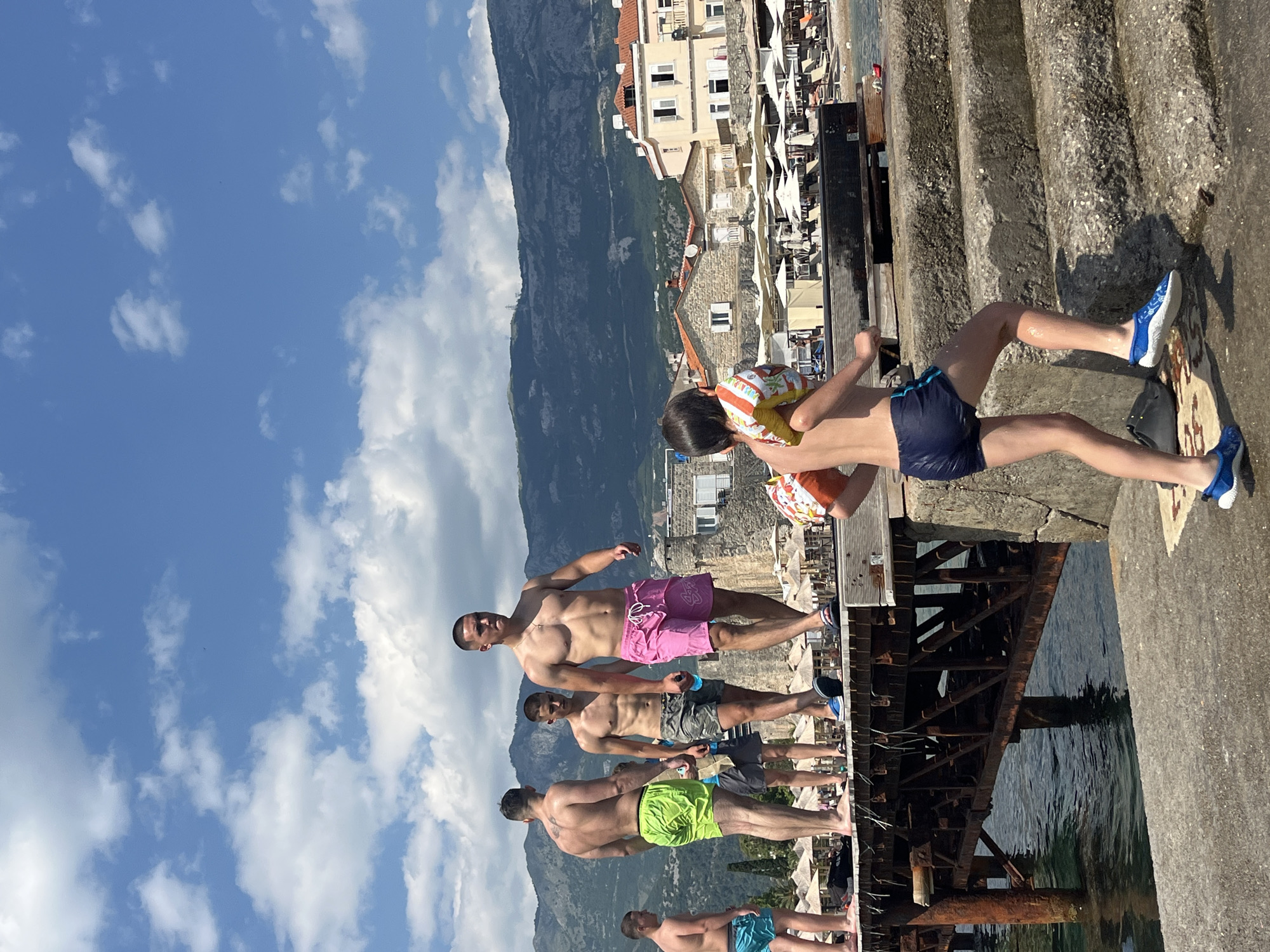 The image depicts a sunny day at a beach or pier area, likely in Budva, Montenegro, given the architectural style of the buildings and the mountainous backdrop.
In the foreground, a young boy with a life vest attached to his upper body is walking up a set of concrete stairs. He is wearing dark blue swimming shorts and blue and white patterned water shoes.
On a wooden pier to the left, five young men are visible. The closest man on the pier is walking towards the right side of the frame and is wearing pink swimming shorts. To his left, another man is walking towards the right and is wearing camouflage patterned swimming shorts. Further to the left, a man in bright green swimming shorts is standing and looking towards the left. Two more men are partially visible on the pier, one wearing turquoise swimming shorts and another in the background. The pier extends over clear water.
In the background, there is a beach with numerous white umbrellas and lounge chairs. Behind the beach, buildings with red-tiled roofs are situated against a backdrop of lush green mountains. The sky is blue with scattered white clouds. The overall scene suggests a recreational summer day at the coast.