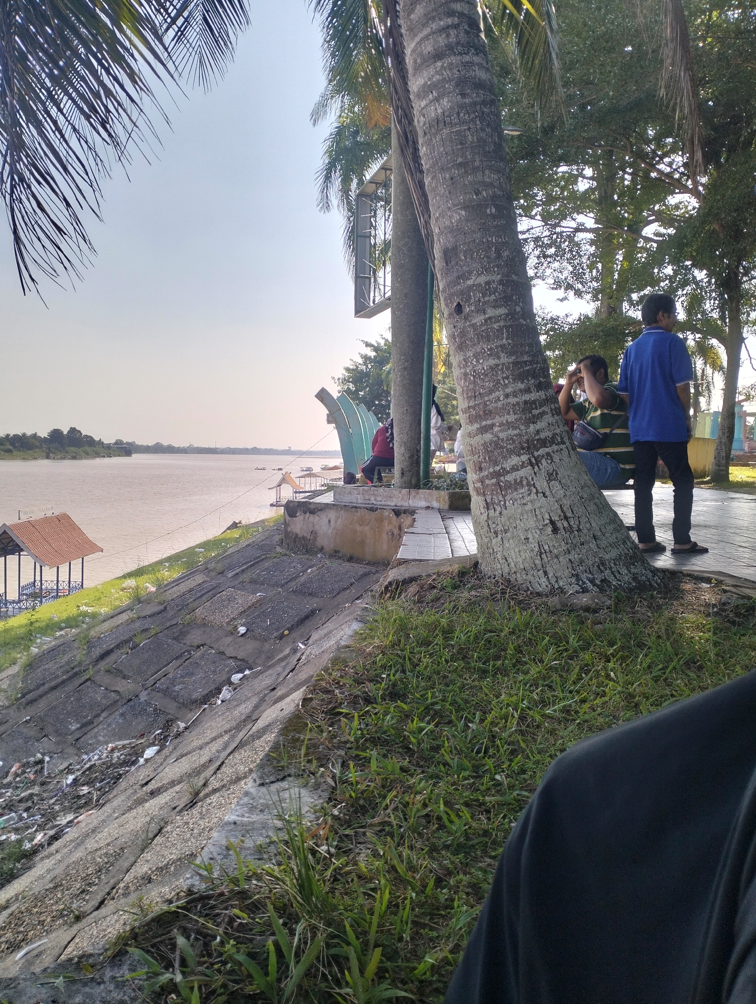 The image depicts a scenic riverside view in Jambi, Indonesia, likely within a park or recreational area adjacent to the Batanghari River. In the foreground, the viewer's shoulder is visible, suggesting the photo is taken from a first-person perspective. The middle ground features a large palm tree trunk with textured bark, leaning prominently into the frame. To the right of the tree, a young boy in a blue t-shirt and black pants stands looking away from the camera. Behind him, a person in a green and yellow striped shirt is seated, leaning against the tree and holding a camera to their face. The background showcases the wide, calm Batanghari River, with a small wooden structure resembling a gazebo or shelter on the left bank. Several small boats and floating platforms are visible on the water. The riverbank is lined with a concrete embankment covered in patches of grass and some scattered debris. Beyond the river, the distant treeline of the opposite bank is visible under a clear, bright sky. The overall scene conveys a peaceful, outdoor atmosphere.