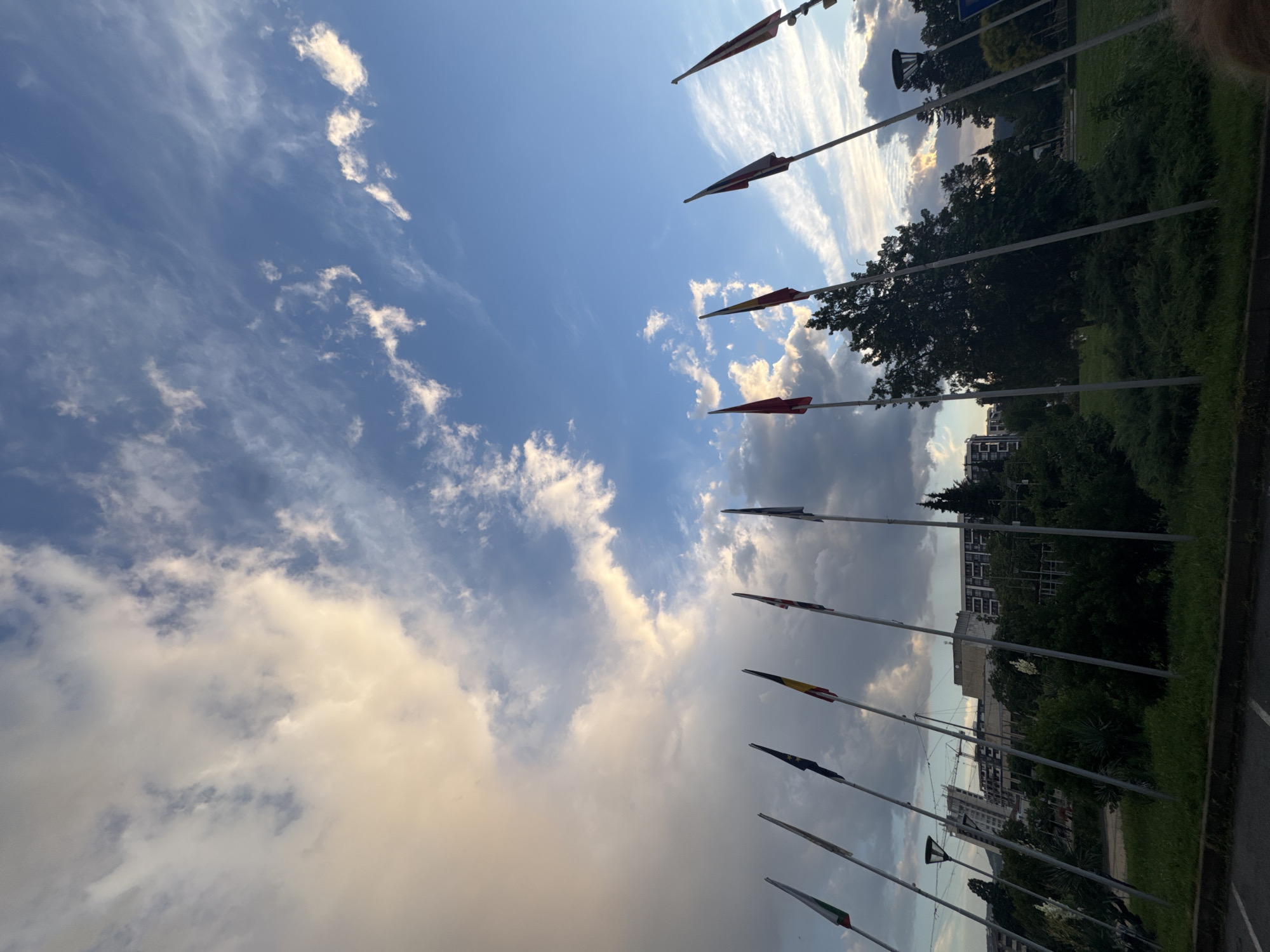 The image depicts a low-angle view of a row of flagpoles with flags flying, set against a dramatic sky with a mix of blue sky and dramatic clouds, some of which are illuminated by sunlight. In the background, there are buildings, trees, and greenery, suggesting an outdoor urban or park setting. The flags are of various designs and colors, and they are arranged in a line, extending from the left to the right of the frame. The overall impression is one of a grand or formal occasion, possibly an international event or a national celebration, given the presence of multiple flags. The lighting suggests it is either early morning or late afternoon.