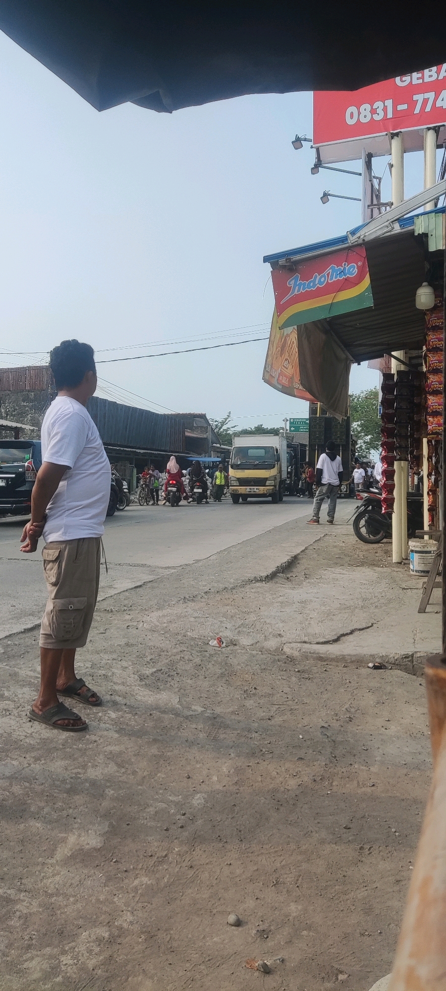 In Gebang, Indonesia, a man wearing a white t-shirt and khaki cargo shorts stands facing away from the camera, looking towards a busy intersection. The ground is rough and uneven, with patches of cracked concrete and dirt. Across the street, a yellow truck is stopped at the intersection. Several motorcycles and people are also visible, some on motorcycles and others walking. Above the intersection, a red billboard with white text, likely advertising, is visible. A smaller sign with the "Indomie" logo hangs from an awning over a shop or stall on the right side of the image. The sky is clear and bright.